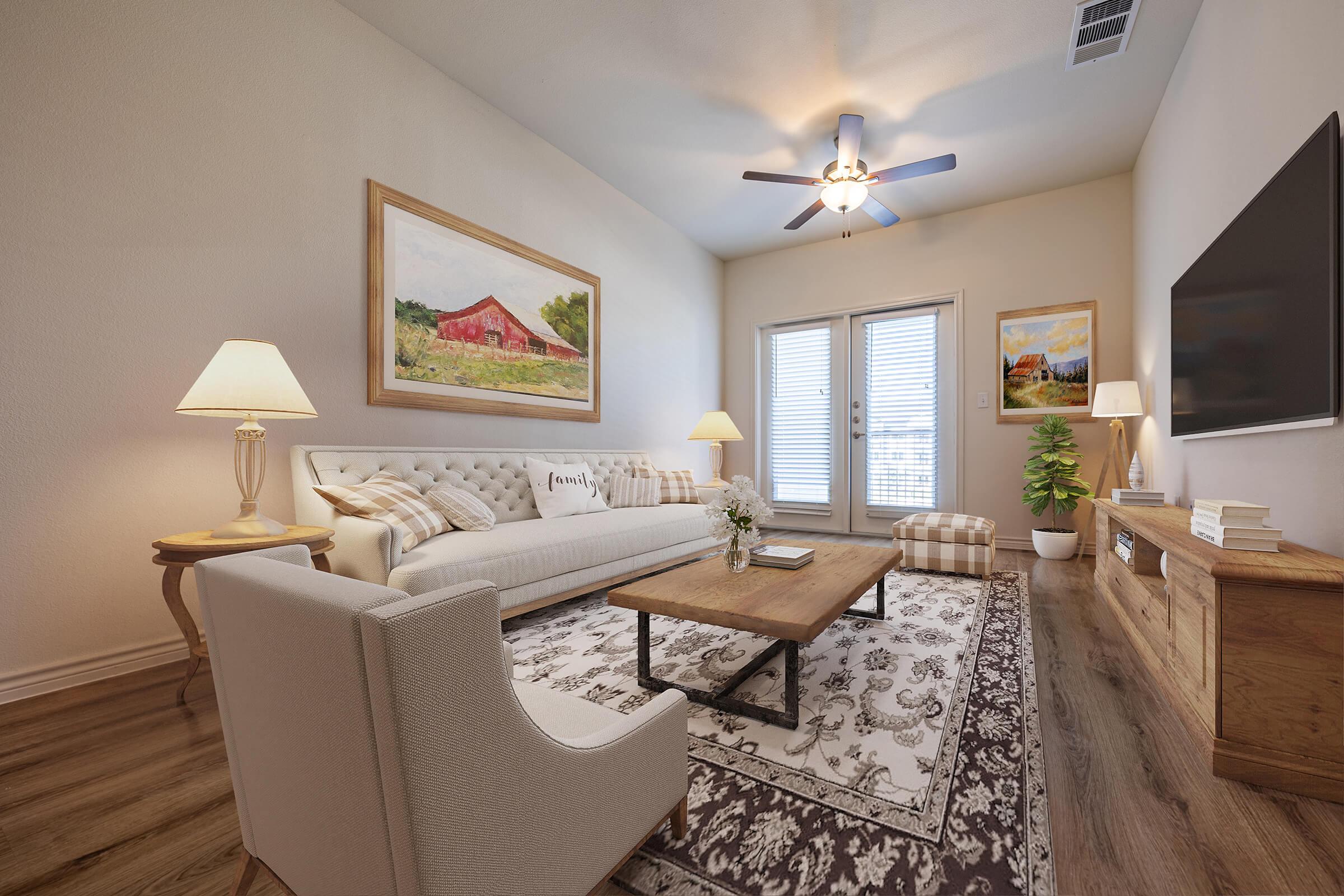 A cozy living room featuring a light-colored sofa with patterned cushions, a wooden coffee table, and a decorative rug. There are lamps providing warm lighting, a flat-screen TV on a wooden console, and artwork on the walls, including a landscape painting. Large windows let in natural light.