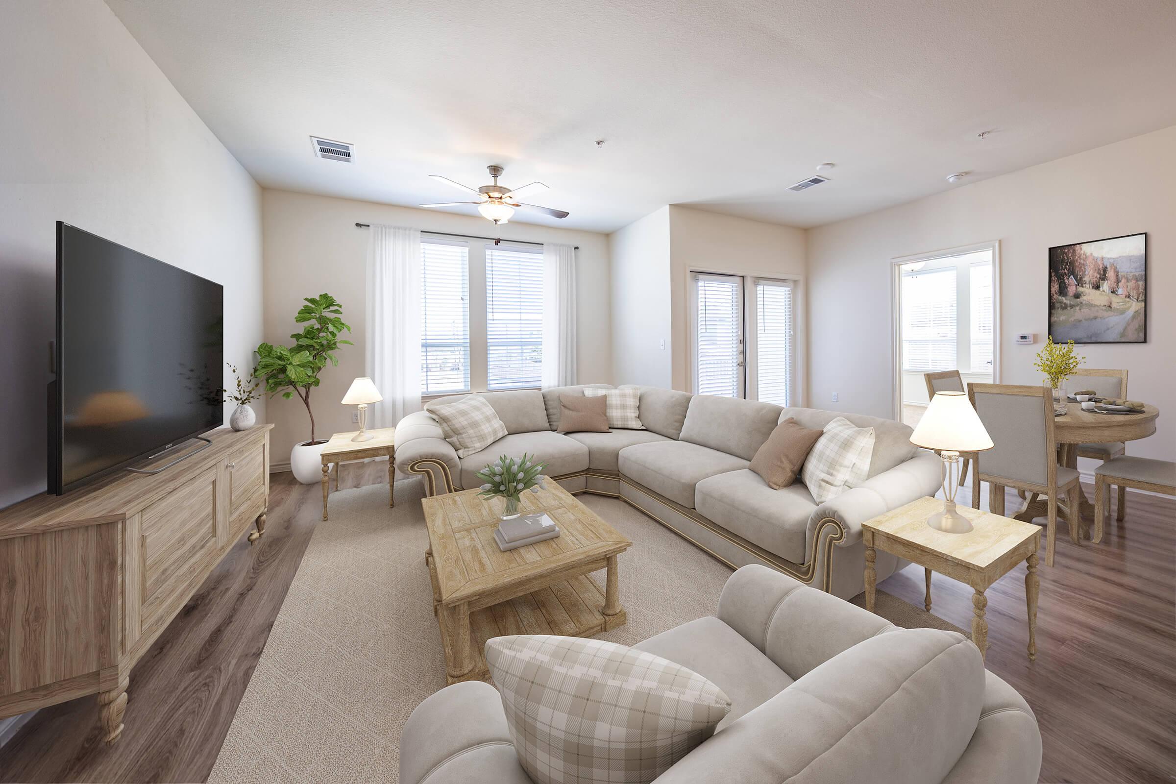 A spacious, modern living room featuring a light-colored sectional sofa, a wooden coffee table, and a TV cabinet. Natural light fills the room through large windows with sheer curtains. A small dining area with a round table and chairs is partially visible. Decorative plants and a table lamp add warmth to the space.
