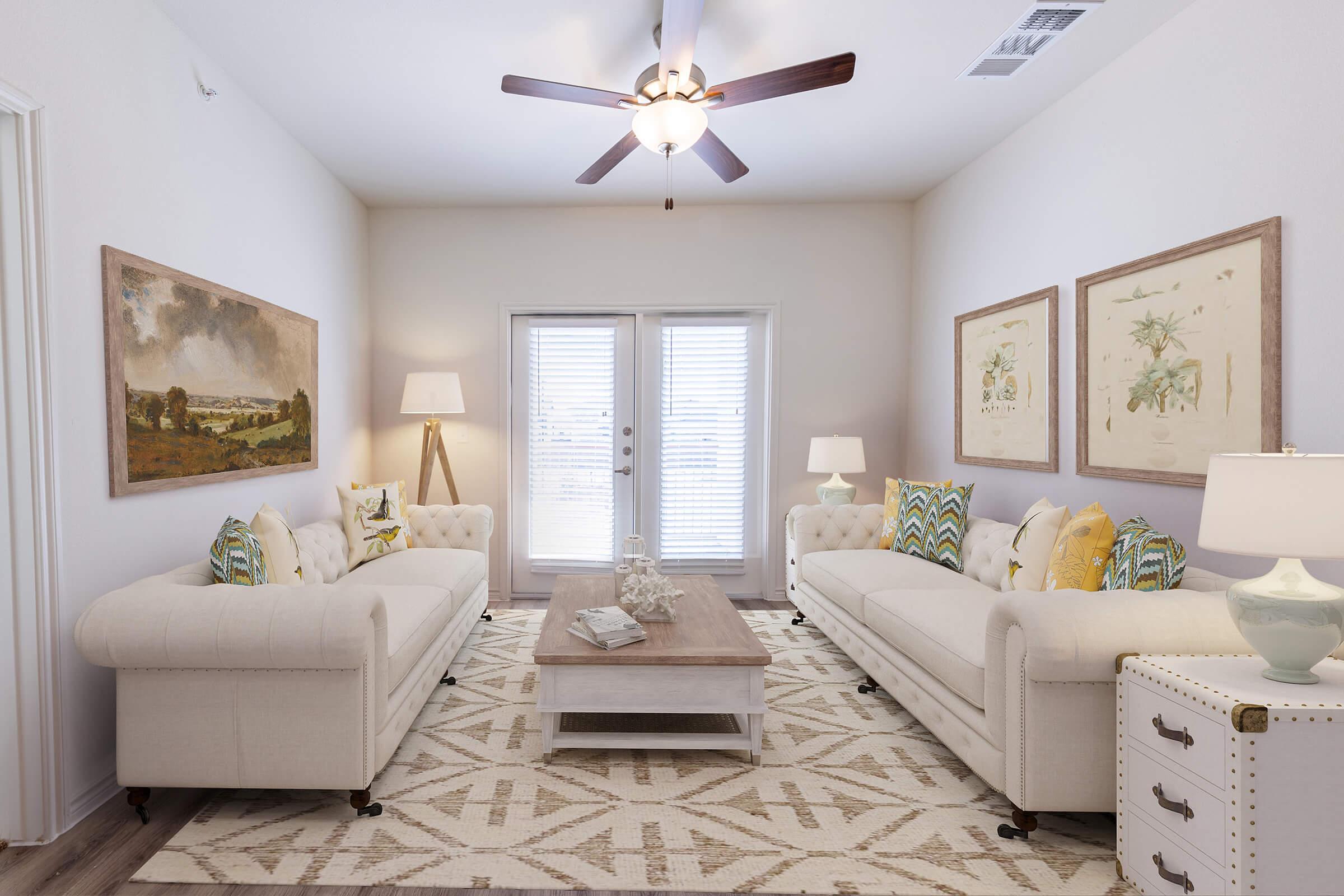 A modern living room featuring two white tufted sofas with decorative pillows, a rustic wooden coffee table, and a neutral area rug. The walls are adorned with framed artwork, and there is a floor lamp and end tables with lamps. Large windows allow natural light to brighten the space.