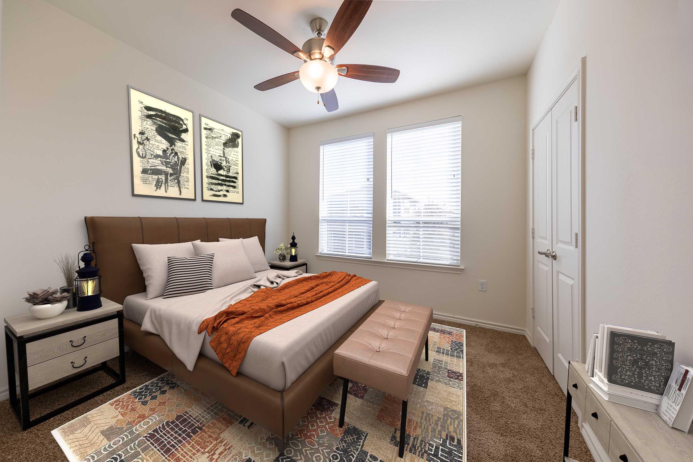 A cozy bedroom featuring a large bed with a brown headboard, topped with white and orange bedding. A bench sits at the foot of the bed. Two framed artworks adorn the walls, and there are windows with blinds allowing natural light. A nightstand with a lamp and decorative items completes the inviting atmosphere.
