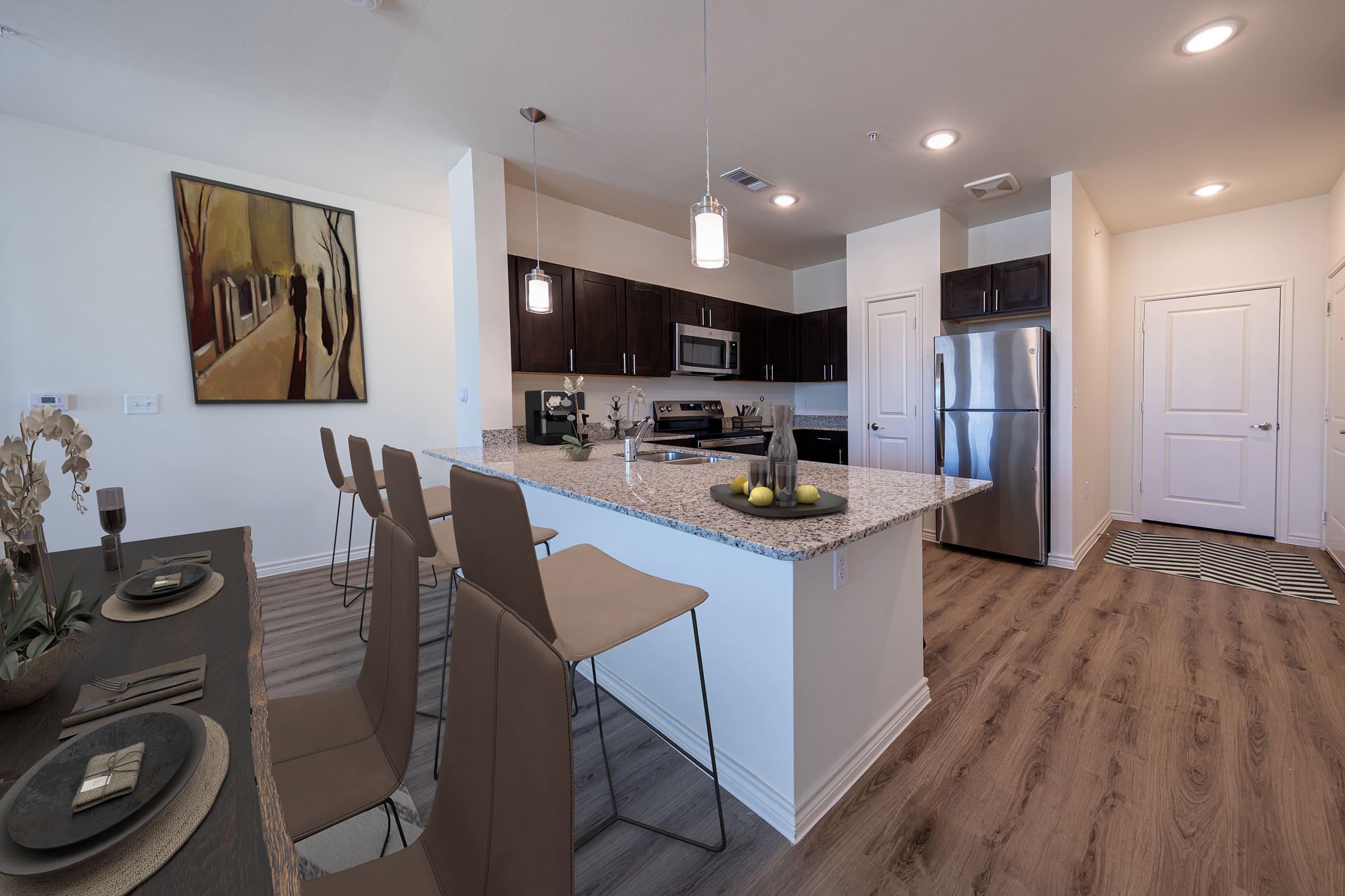 Modern kitchen featuring a large island with bar stools, stainless steel appliances, and granite countertops. A dining table is set on one side, and there's a wall painting of a street scene. Light-colored walls and wooden floors create a welcoming, open atmosphere. A doorway leads to a hallway.