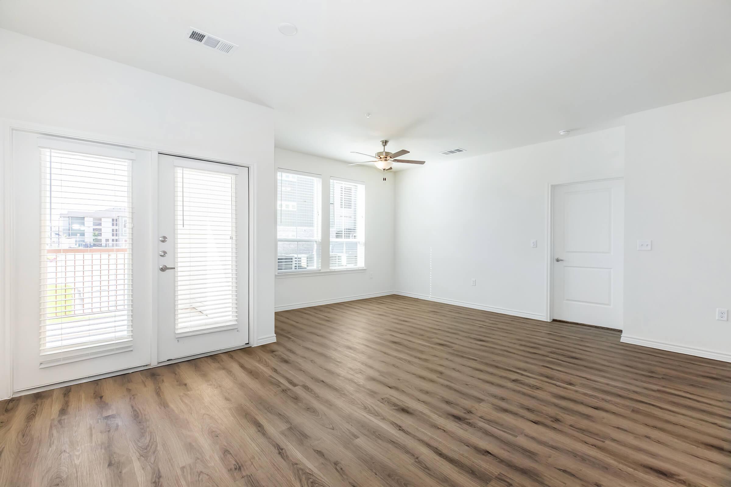 Bright, spacious living area with large windows and light wood flooring. Features a ceiling fan and access to a balcony through glass doors. Modern and minimalistic design with white walls and ample natural light.