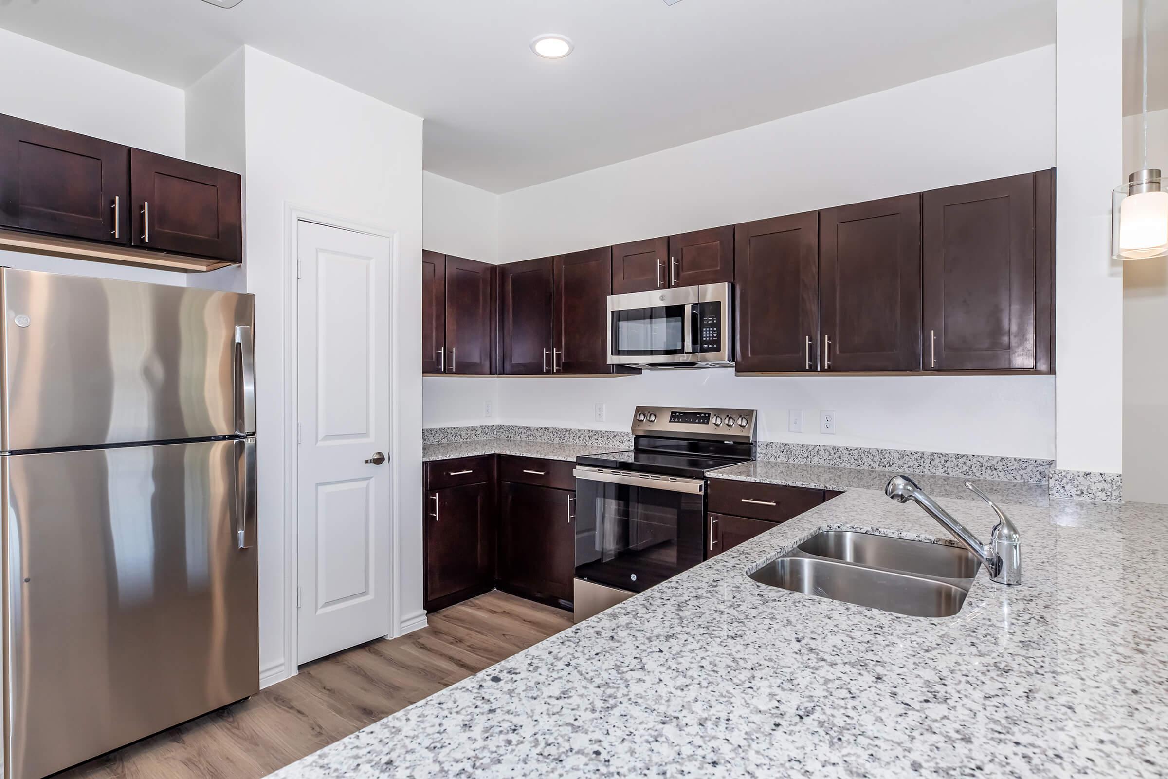 Modern kitchen featuring dark wooden cabinets, stainless steel appliances including a refrigerator and microwave, a granite countertop, a sink, and a stylish light fixture. The space is bright with natural lighting and has a clean, contemporary design.
