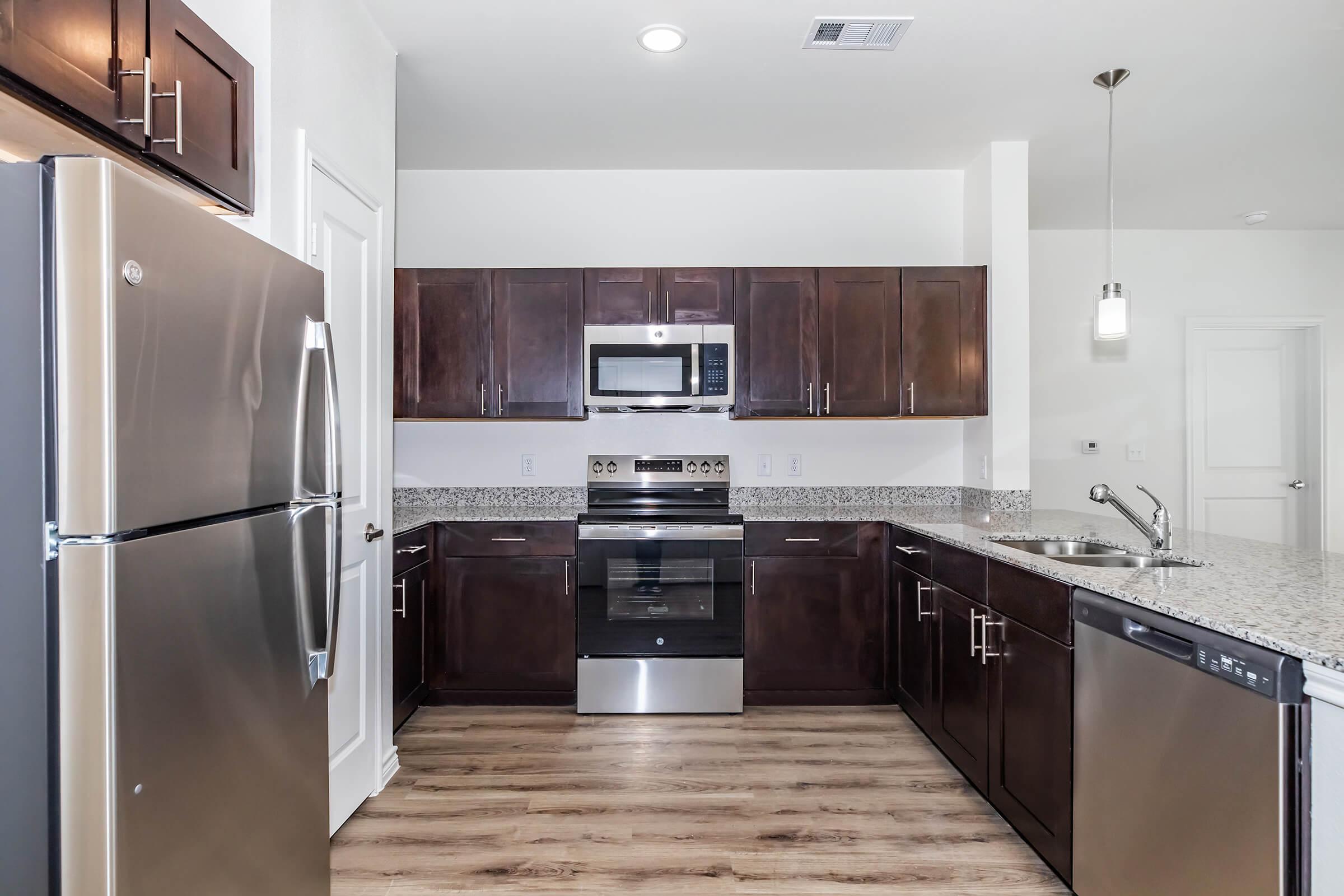 Modern kitchen with stainless steel appliances, including a refrigerator, microwave, oven, and dishwasher. Dark wooden cabinets contrast with the light granite countertops. The space features a sleek design with a pendant light hanging above the sink and a warm wood-like floor.