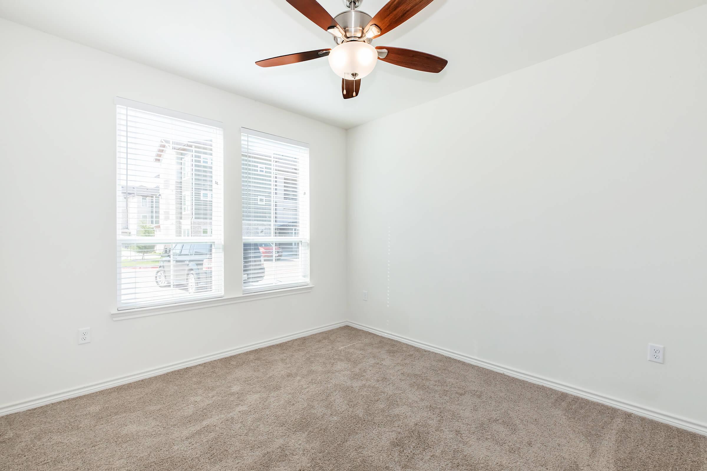 Empty room with light beige carpet and white walls. Two windows with blinds are on one side, allowing natural light to enter. A ceiling fan with wooden blades is centered above. The room appears spacious and well-lit, ideal for personal décor and furnishings.
