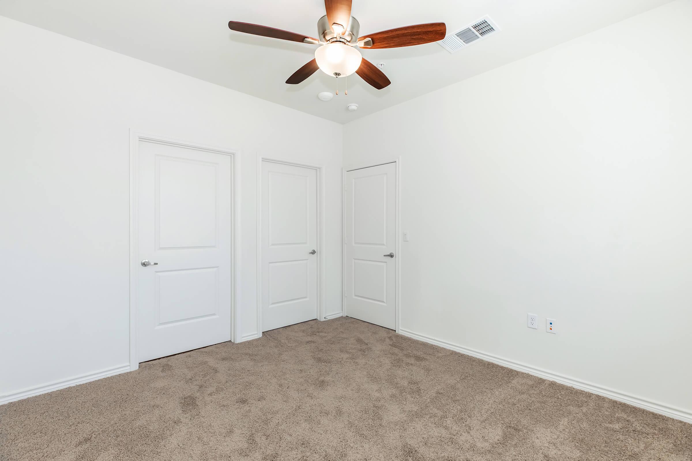 A bright, empty room with light-colored walls and a tan carpet. The room features a ceiling fan with wooden blades and has three white doors on one wall. Natural light fills the space, creating an open and airy atmosphere.