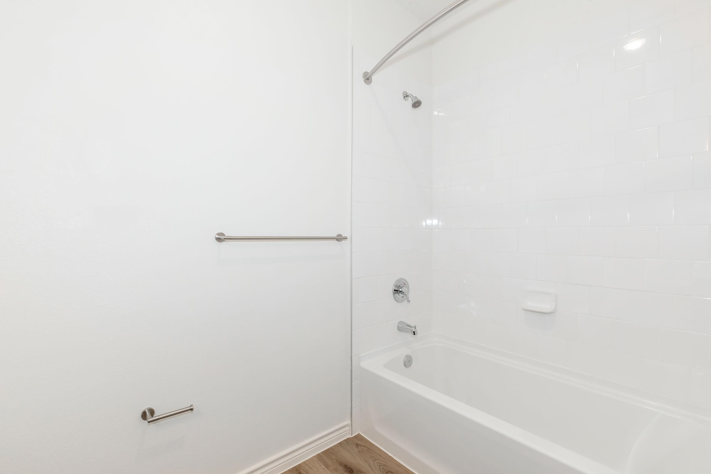 A clean, minimalist bathroom featuring a white tiled wall, a bathtub with a showerhead, and a towel bar. The floor is light-colored wood, enhancing the bright and spacious feel of the room.
