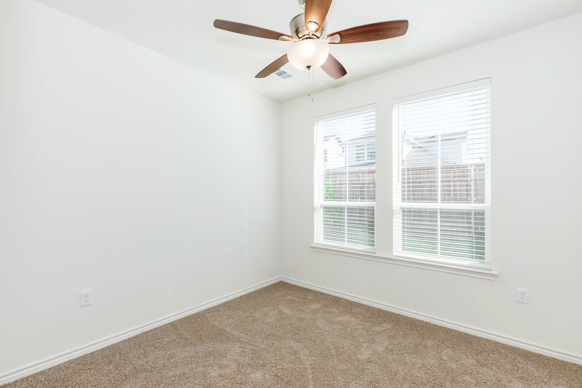 Empty room with light beige carpet, two large windows with white blinds letting in natural light, and a ceiling fan with wooden blades. The walls are painted white, creating a bright and airy atmosphere.