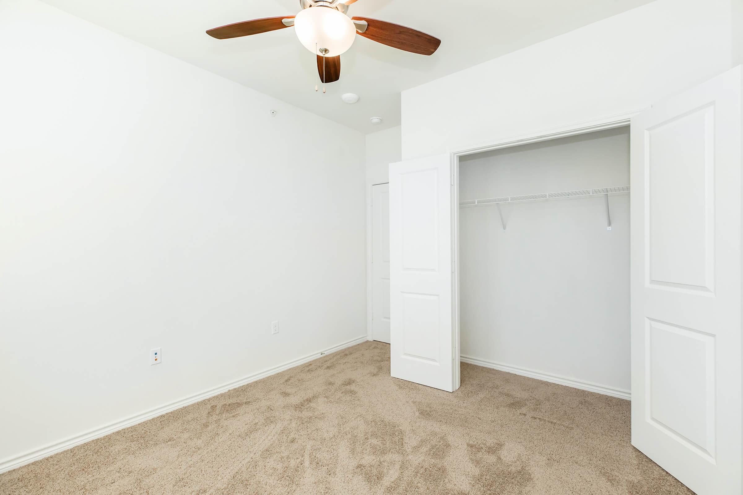 A simple, empty bedroom featuring light-colored walls, beige carpet, and a ceiling fan. A closet with sliding doors is visible on one side, and the room has a bright and airy feel with ample natural light.