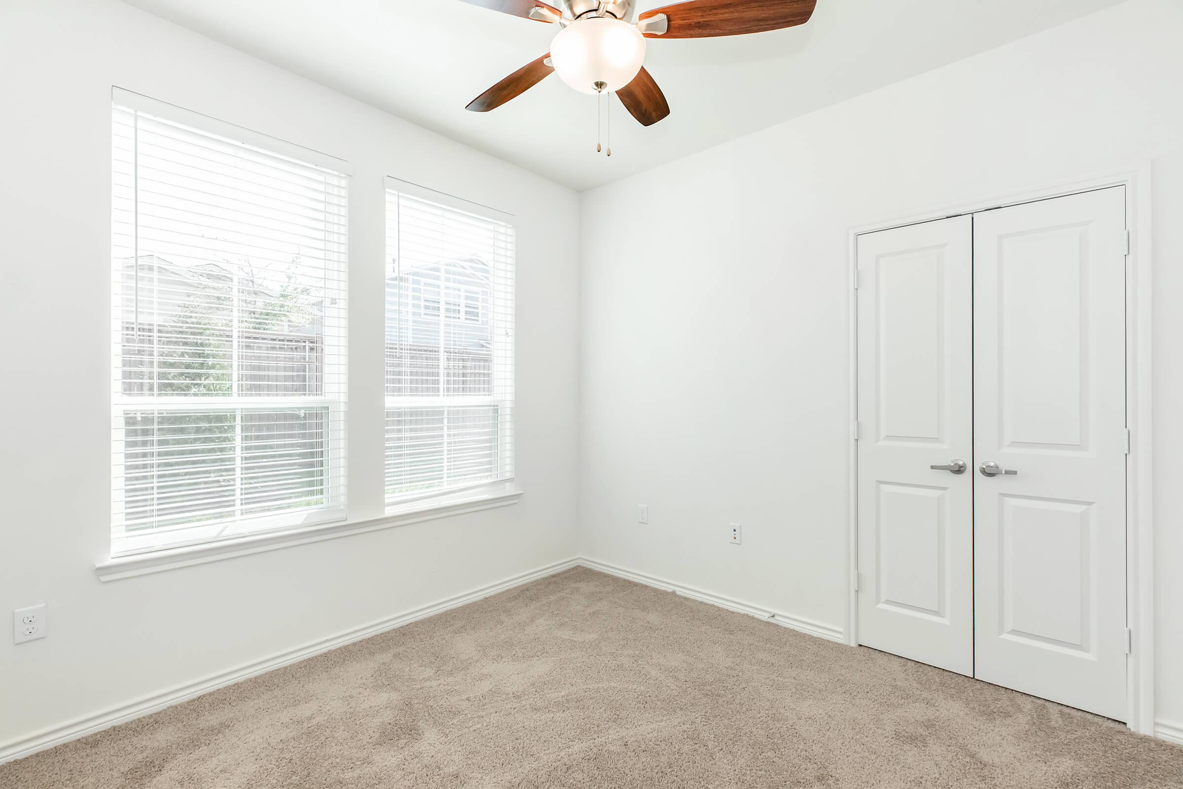 A well-lit, minimalist room featuring two large windows with blinds, a light ceiling fan, and light-colored walls. The floor is carpeted in a neutral tone, and there is a double door closet on one wall. The overall ambiance is clean and inviting, suitable for various purposes.