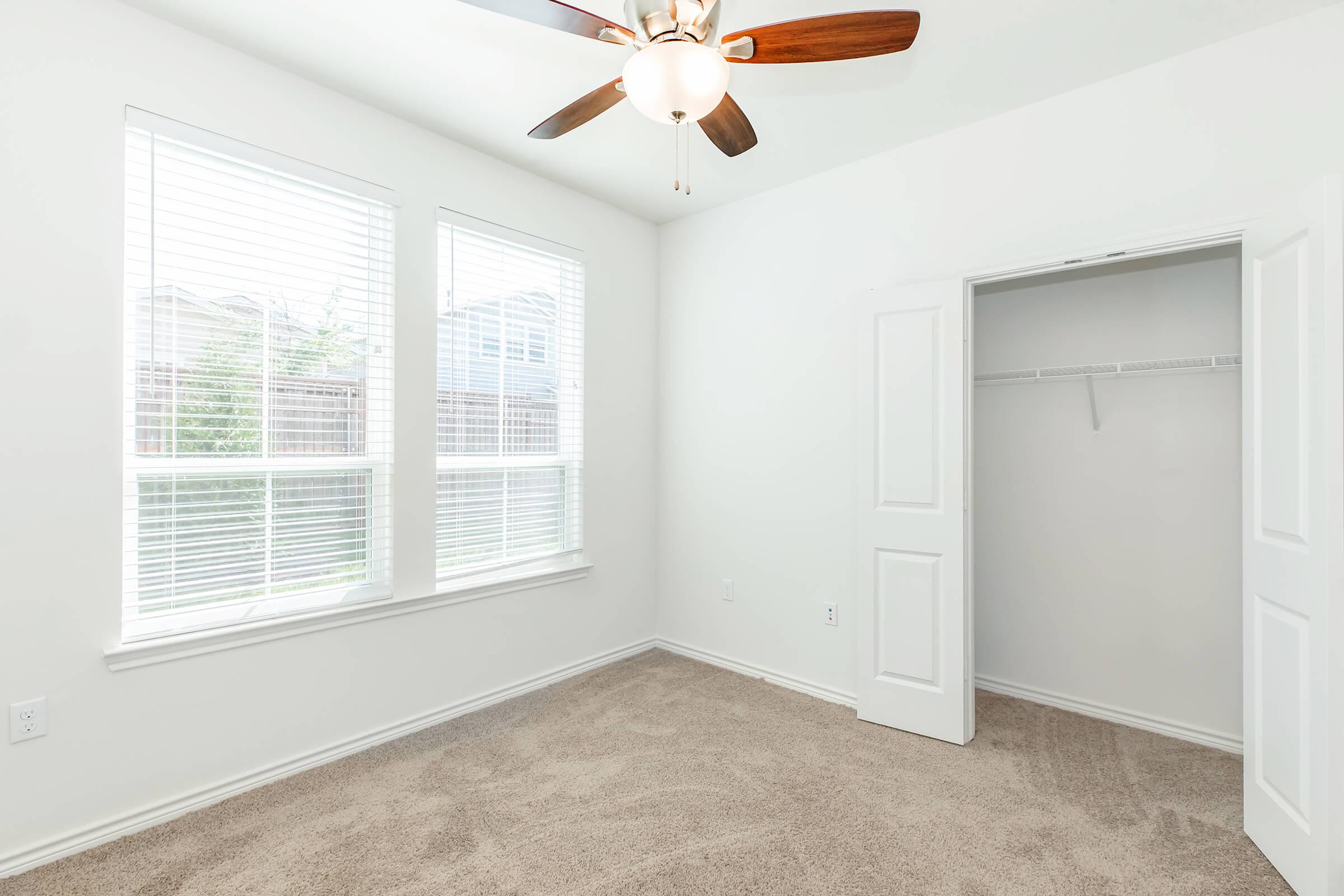 A well-lit empty bedroom featuring two windows with blinds, a ceiling fan, and beige carpet. One side of the room has an open closet with sliding doors, while the walls are painted white, creating a bright and airy atmosphere.