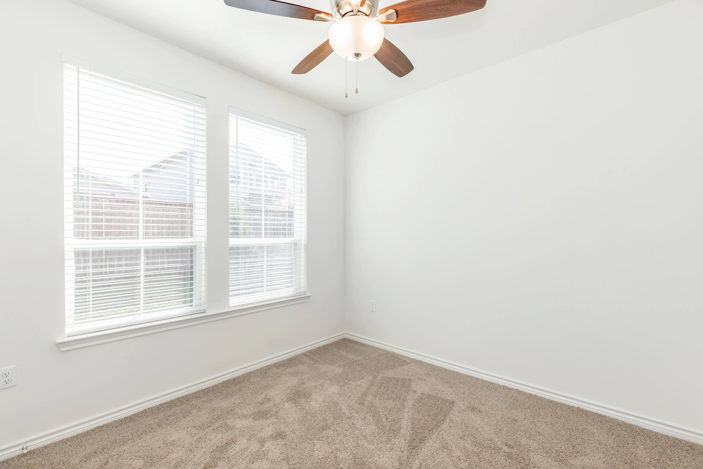 Empty room with light beige carpet, white walls, and two large windows with white blinds. A ceiling fan with wooden blades hangs from the ceiling, providing a well-lit, spacious atmosphere. The room is uncluttered, ideal for various uses.