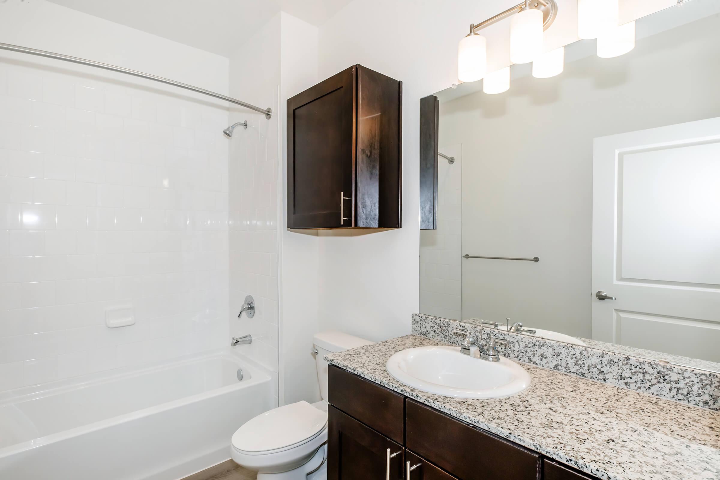 Modern bathroom featuring a white tiled bathtub with a shower, dark wood cabinetry, a granite countertop with an oval sink, and a large mirror above the sink. Soft lighting is provided by three wall-mounted sconces. A door leads to another room, and the overall aesthetic is clean and minimalist.