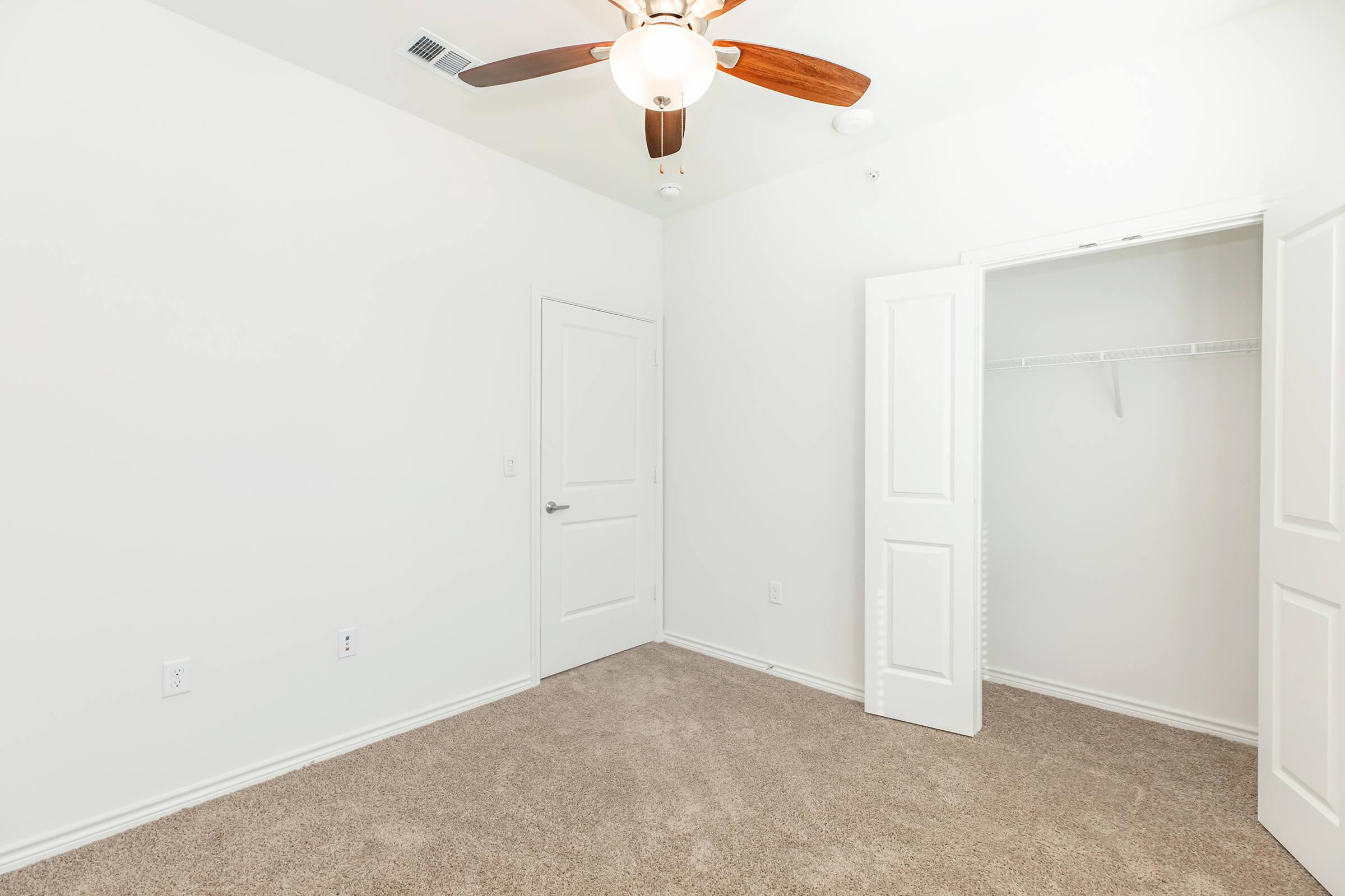 A clean, empty room featuring light-colored walls and carpet. There is a ceiling fan with wooden blades and a closet with white doors. Natural light fills the space, creating a bright and airy atmosphere.