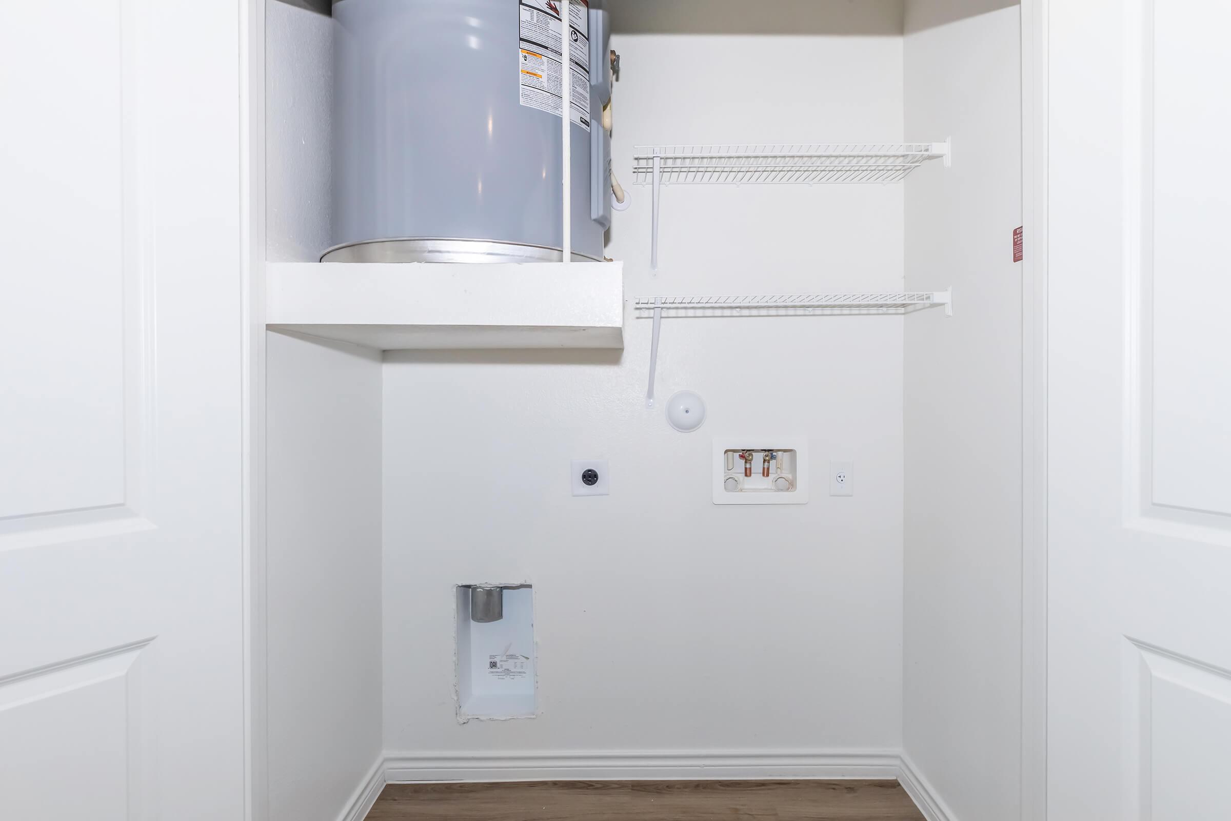 A small laundry room with a water heater, wire shelves, and wall outlets. The walls are painted white, and the floor has a light wood finish. There is a rectangular cutout in the wall, and a round sensor or device is mounted nearby. The overall space is tidy and functional for laundry tasks.