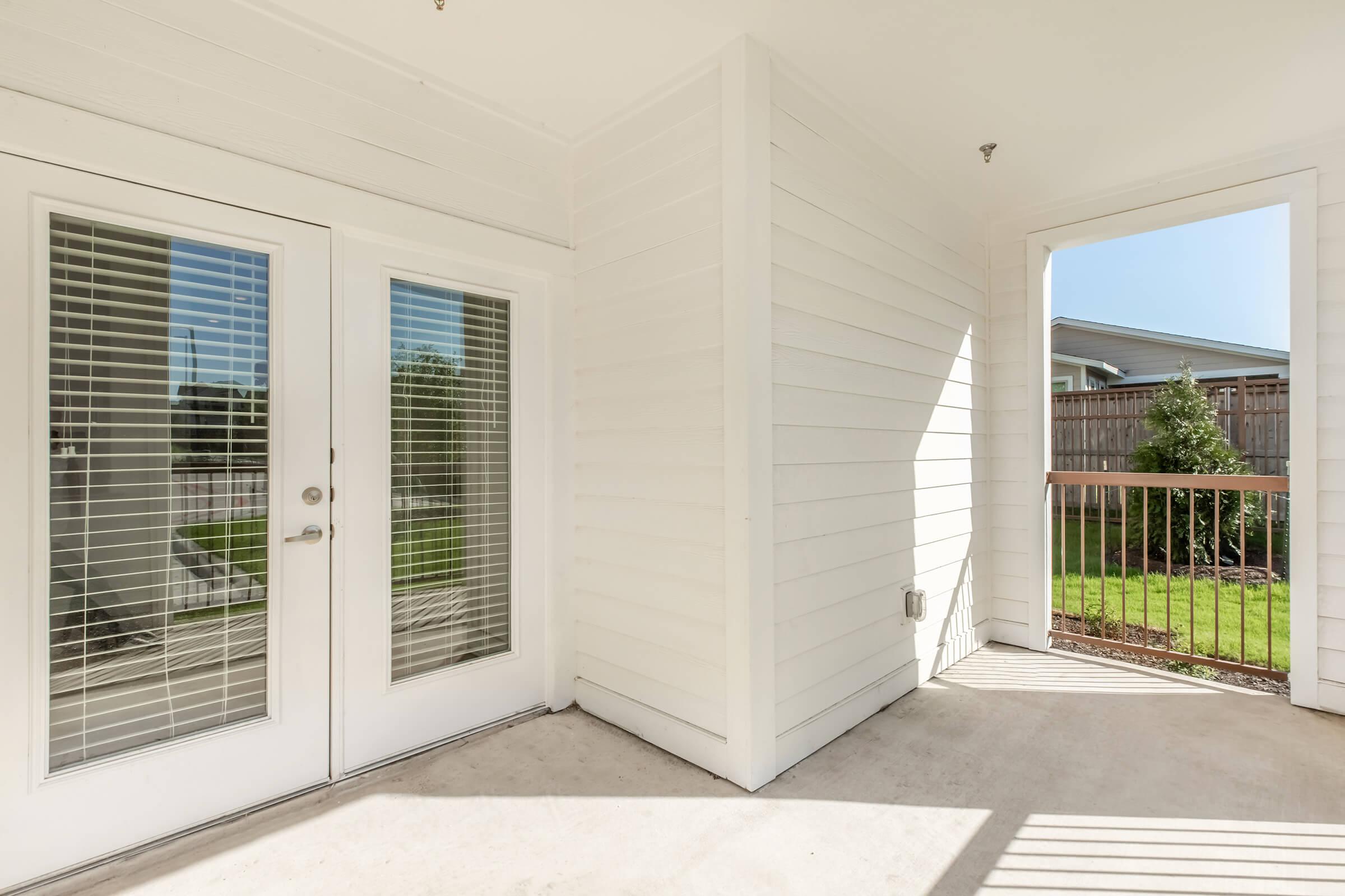 Bright and airy outdoor space featuring a covered patio with white wooden siding, double glass doors with blinds, and an open area leading to a balcony. Lush green grass is visible through the doors, with a wooden fence in the background. Ideal for relaxation or entertaining.