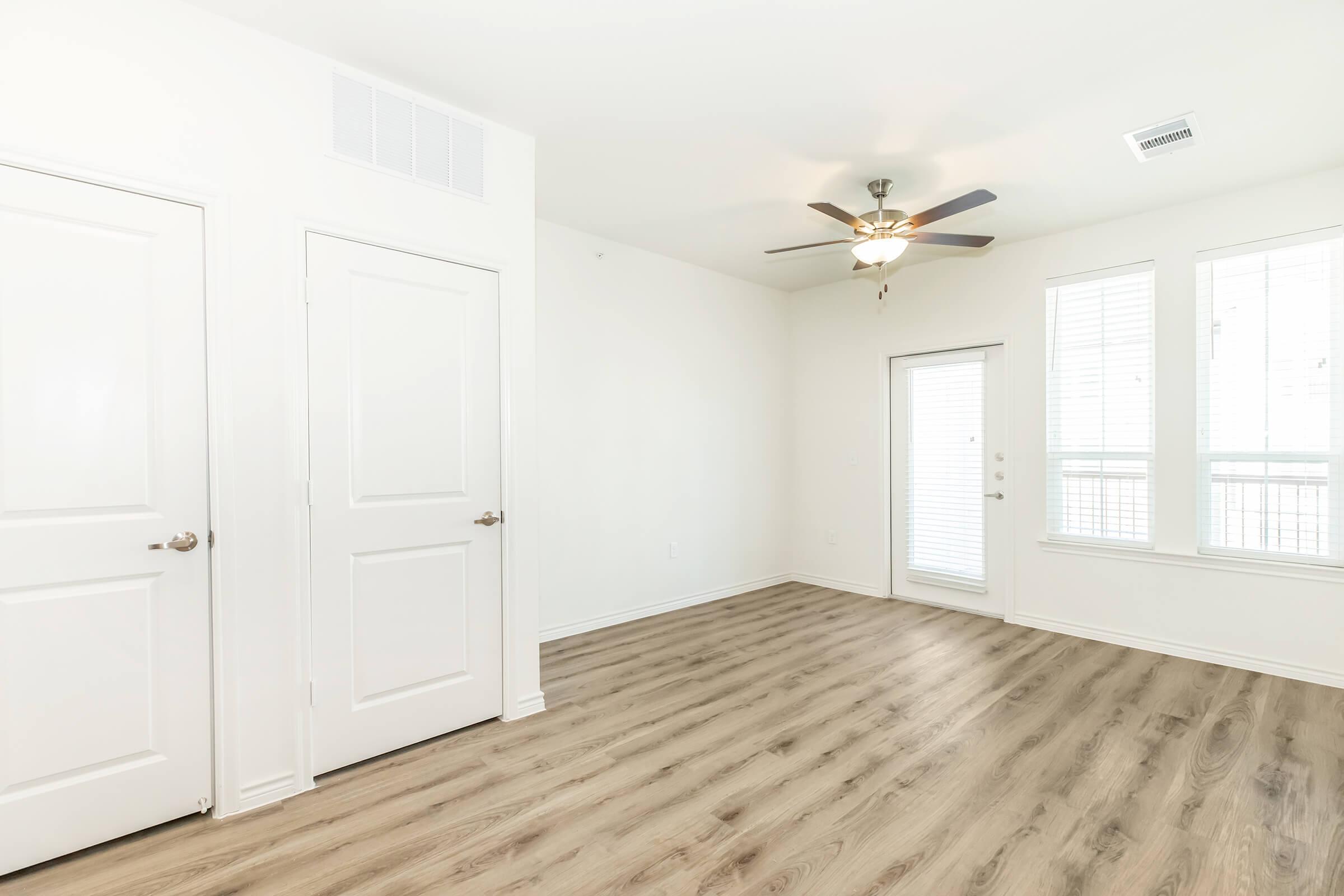 A spacious, modern room with light-colored walls and wooden flooring. There are two closed doors on the left, a sliding glass door leading outside on the right, and large windows allowing natural light. A ceiling fan is visible in the center of the room. The overall ambiance is bright and inviting.