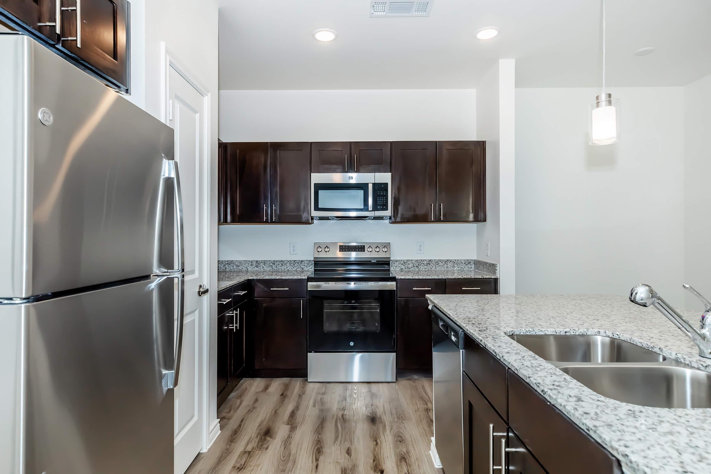 Modern kitchen featuring dark wood cabinetry, stainless steel appliances including a refrigerator and microwave, and a granite countertop. The space has an open layout with a view toward the sink and an island, illuminated by pendant lighting, and a light-colored floor adds warmth to the design.