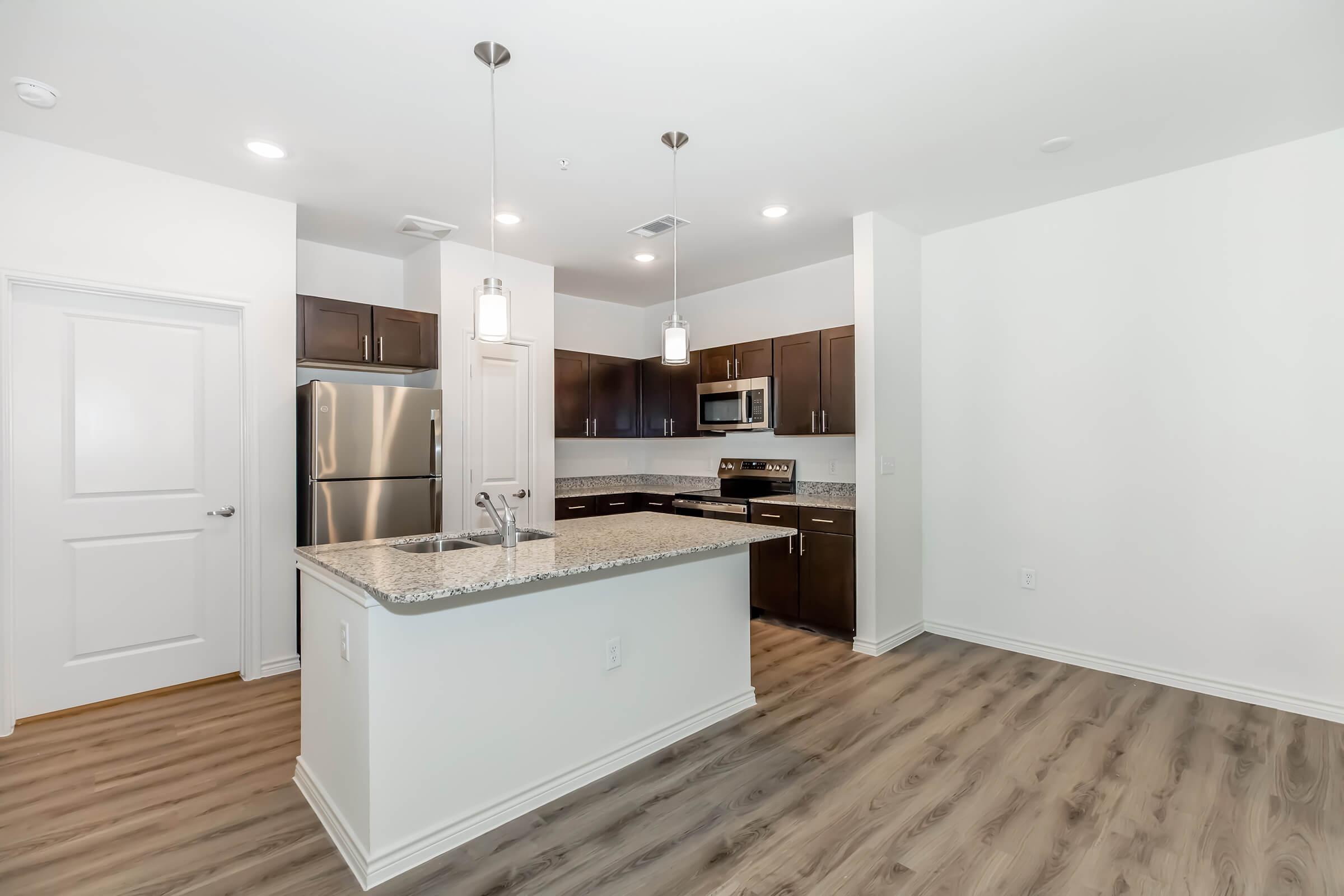 Modern kitchen in an open layout featuring dark wood cabinets, stainless steel appliances, and a large granite-topped island. Bright lighting from pendant fixtures and ample natural light enhance the space, which has warm, wood-like flooring and white walls.