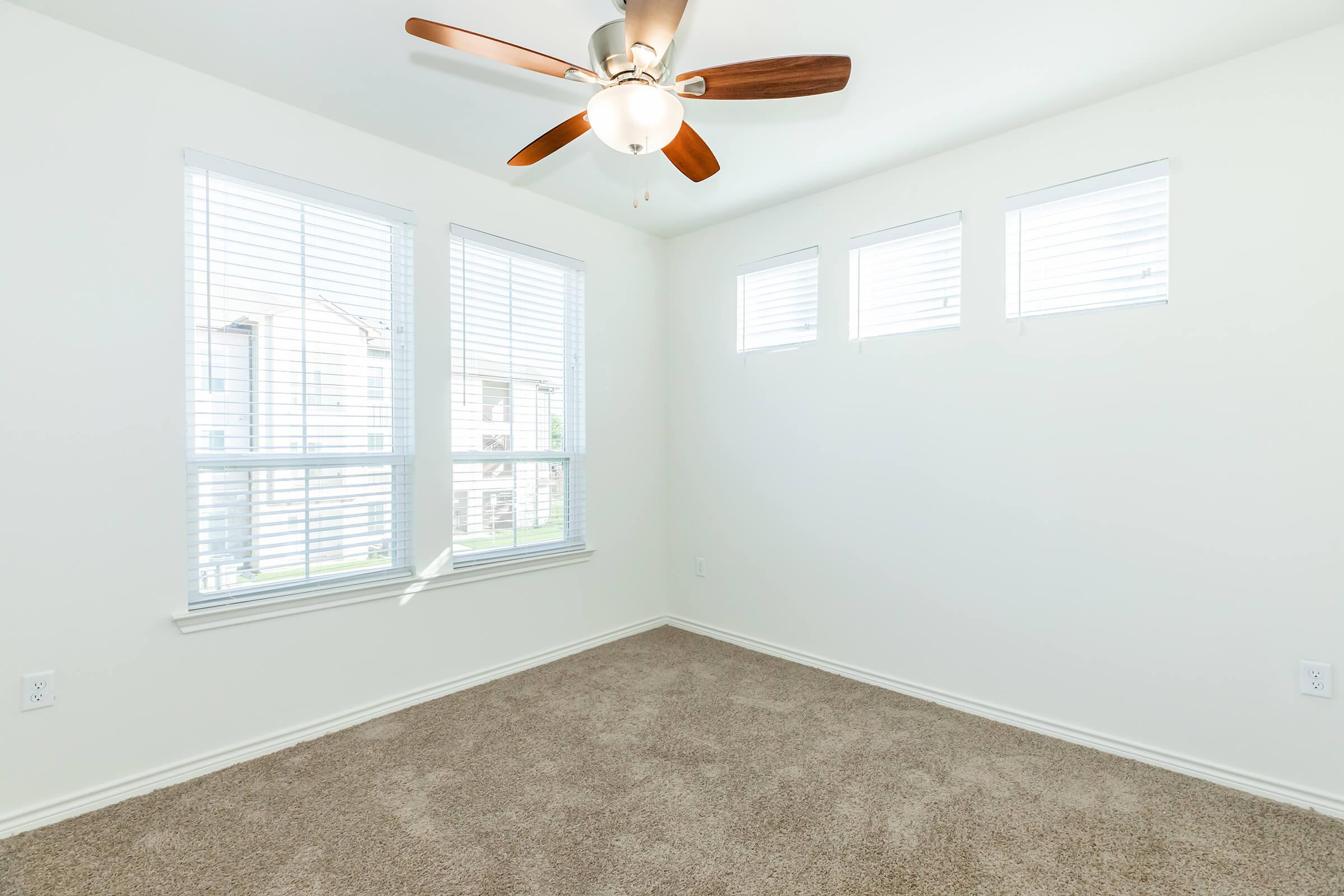 A well-lit empty room featuring light beige carpet and white walls. It has a ceiling fan with wooden blades and three small windows with white blinds, allowing natural light to fill the space. The room is clean and unfurnished, creating a blank canvas for personal decoration.