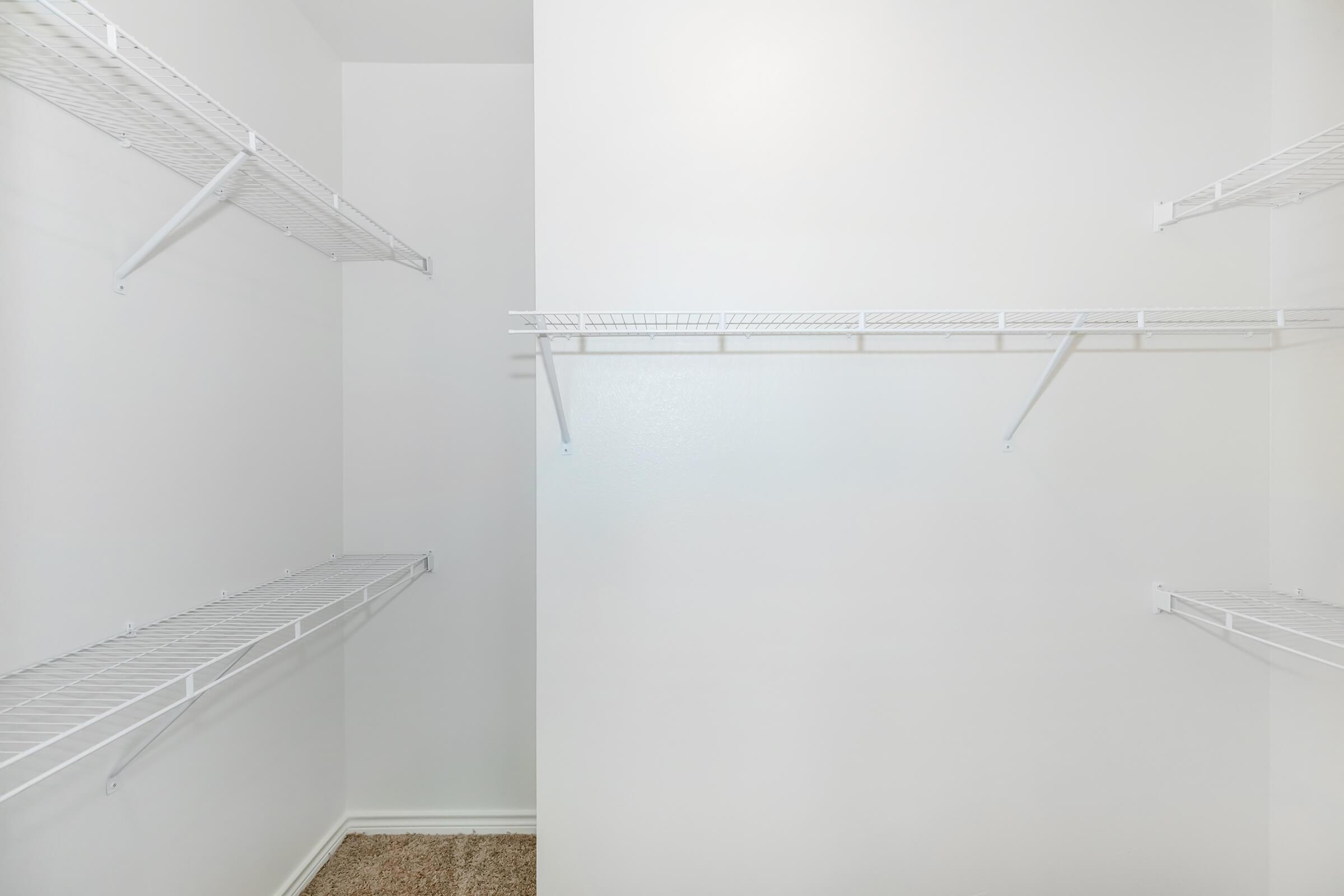Empty closet with white walls and two wire shelving units on the left and one on the right. The floor is carpeted, and the space appears well-lit and clean, providing a minimalistic storage area.