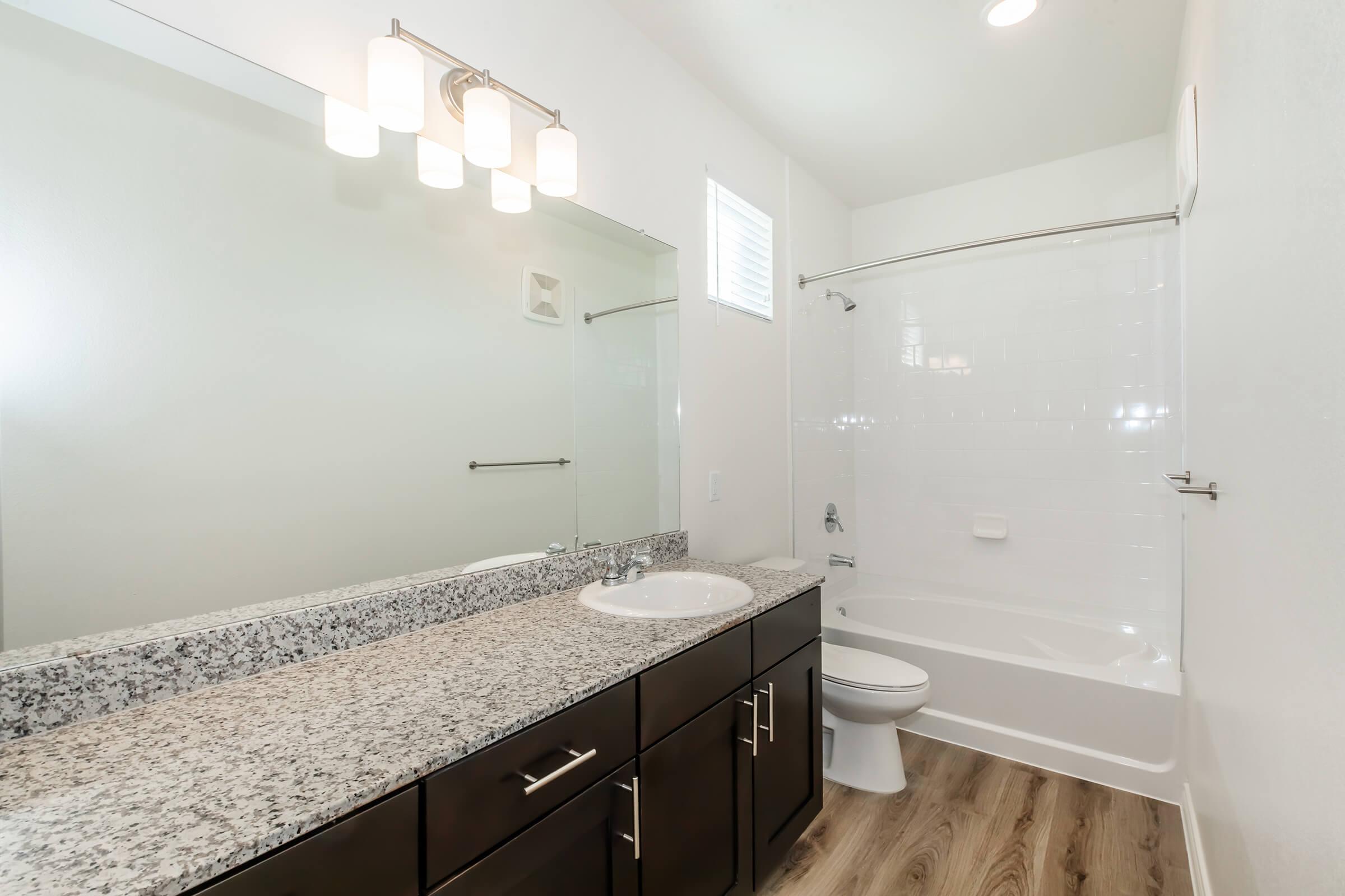 A modern bathroom featuring a granite countertop with a sink, a large mirror with elegant lighting above, a bathtub with a shower, and a toilet. The walls are painted white, and the floor has a wood-like appearance. Natural light comes through a small window.