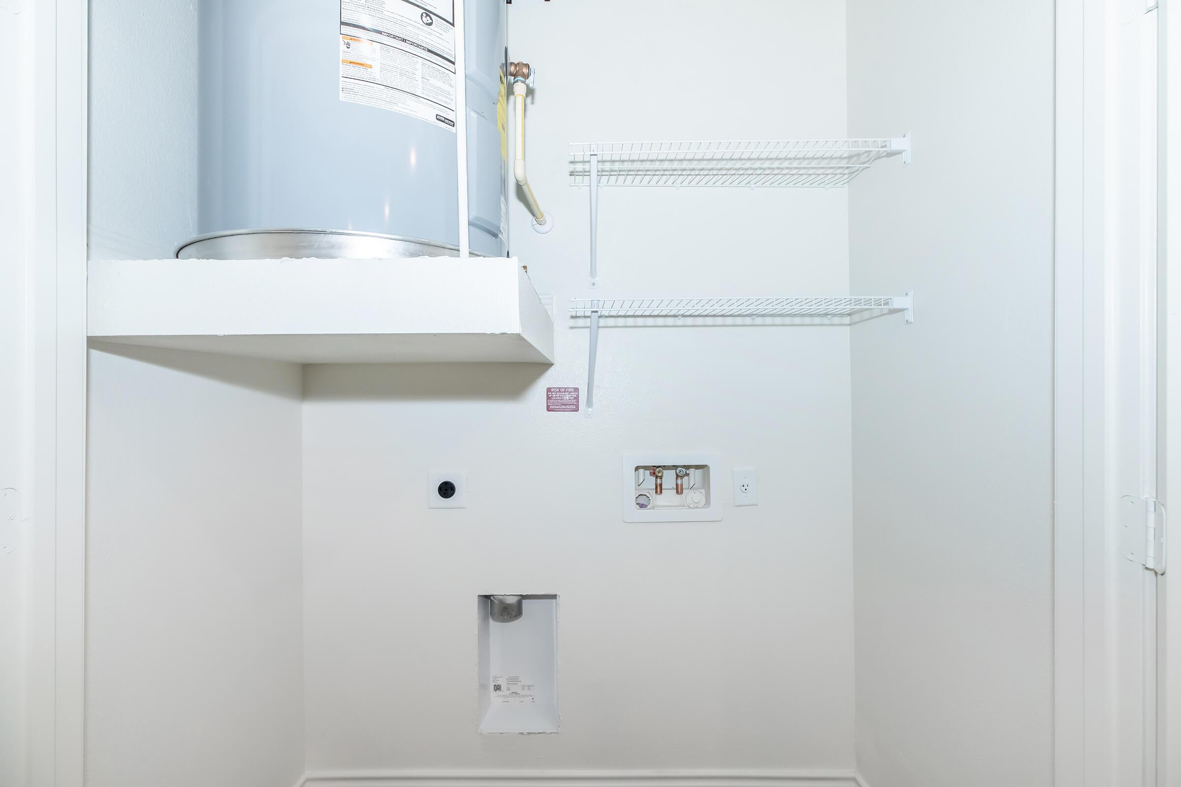 A clean, empty laundry closet featuring a mounted water heater, white shelving on the wall, and an access panel on the lower section. The walls are painted white, and the floor is clear and uncluttered.