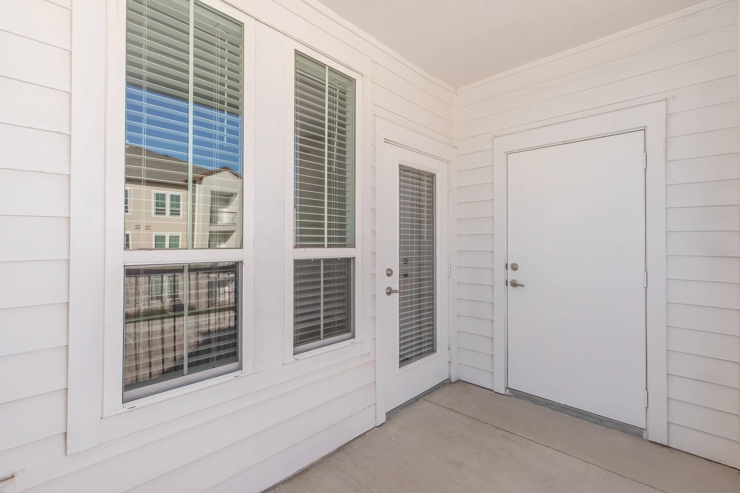 A corner of a porch featuring white wooden siding, two large windows with blinds, and a door alongside a smaller door. There are visible reflections of nearby buildings in the windows. The floor is concrete, and the overall setting is bright and airy.