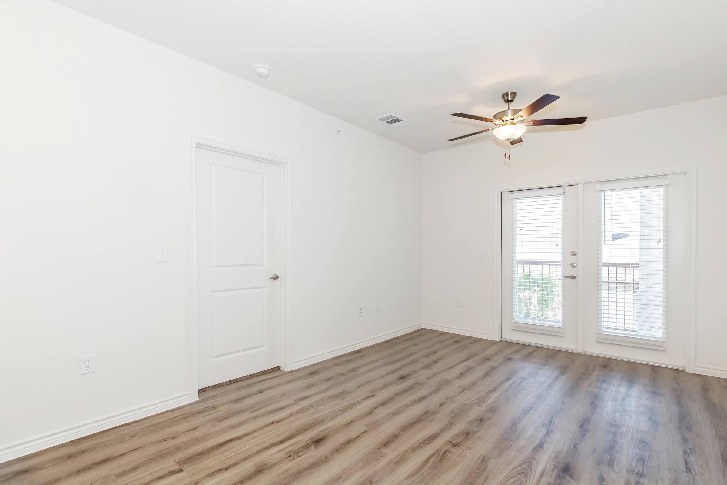 A spacious, well-lit room with light-colored walls and hardwood flooring. There is a ceiling fan for ventilation, a closed door leading to another area, and a pair of sliding glass doors that open to a balcony or patio.