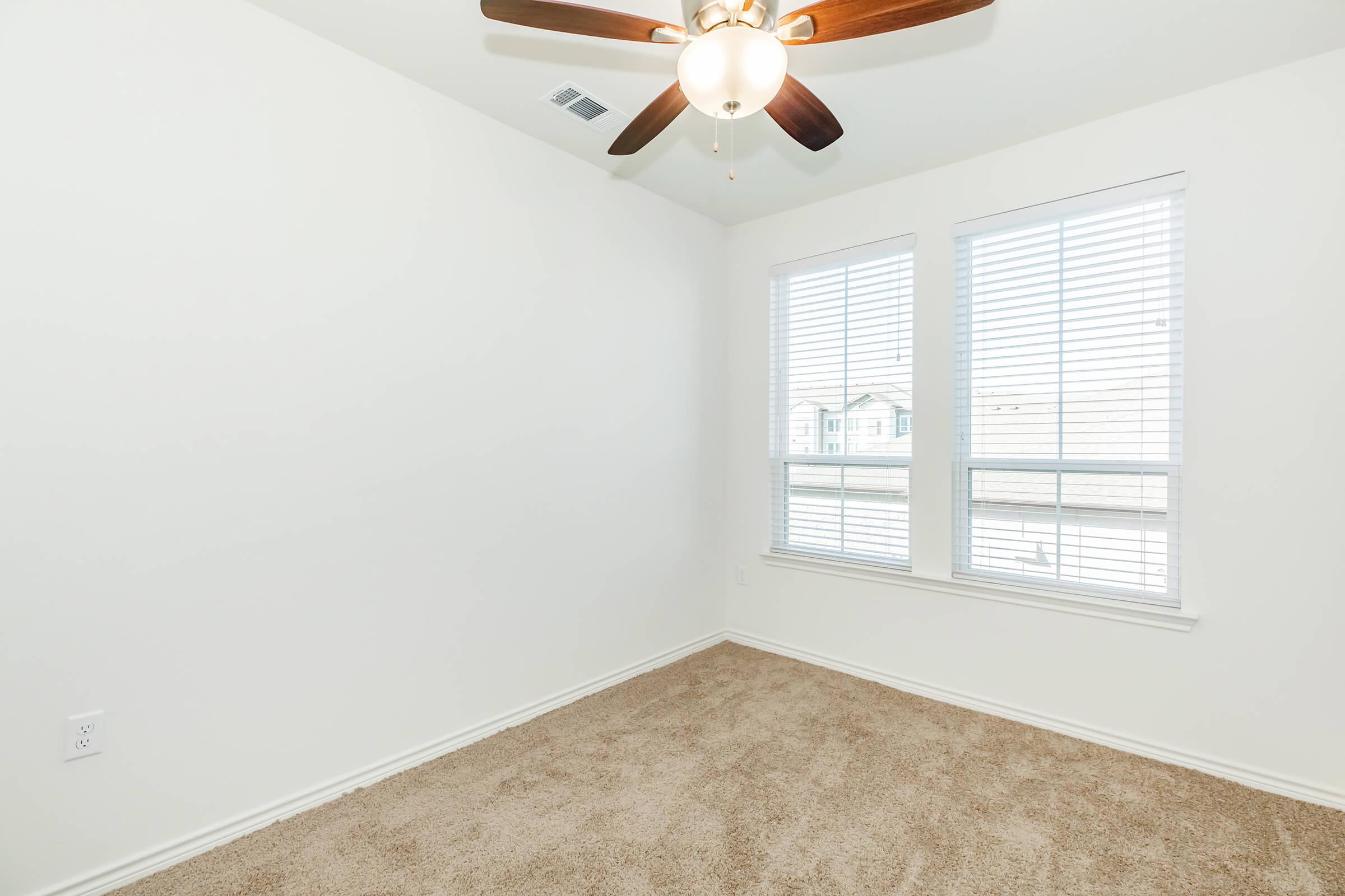 Empty bedroom with light beige carpeting, white walls, and a ceiling fan. Two windows with blinds allow natural light into the room.