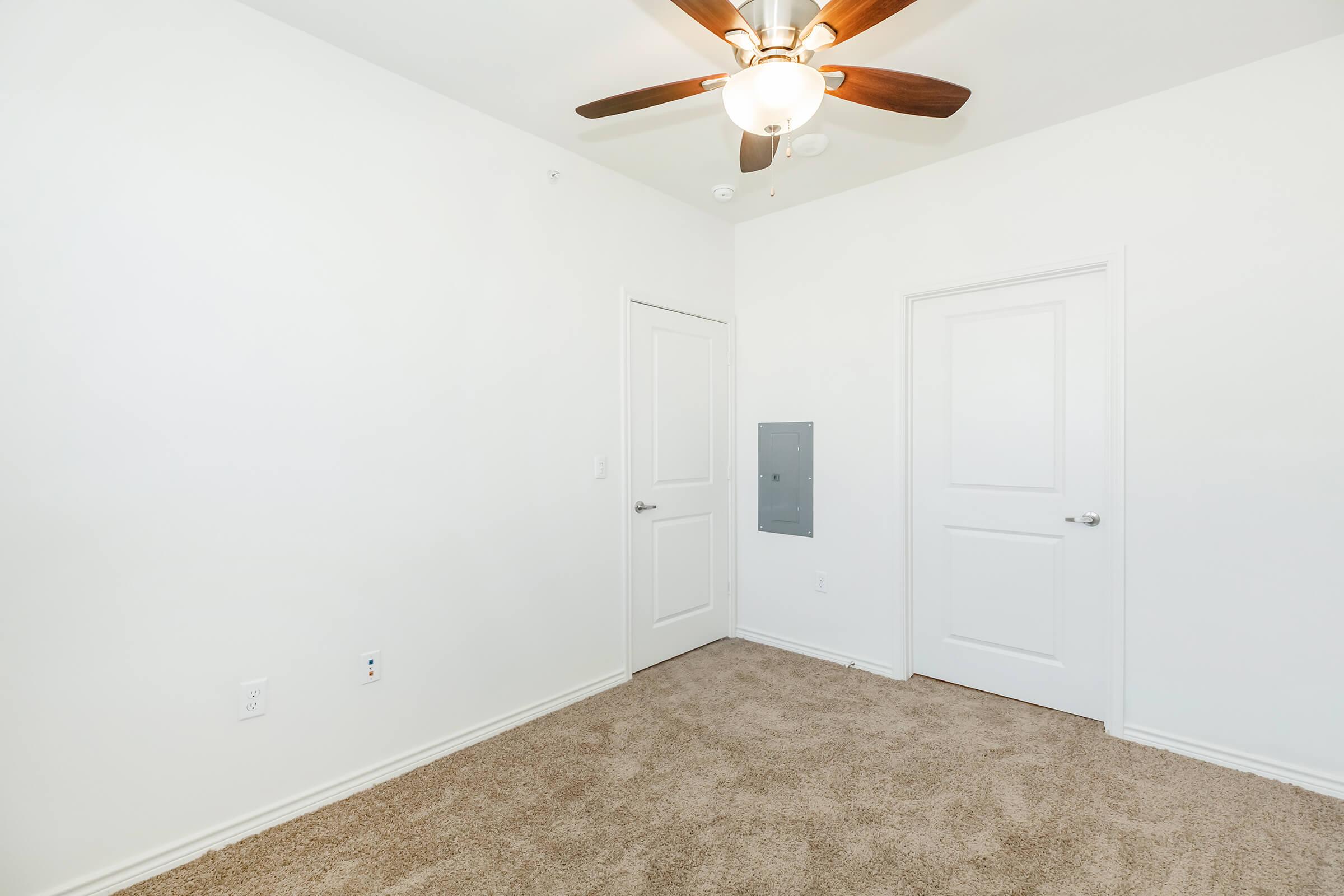 Empty room with light beige carpet, white walls, and a ceiling fan with wooden blades. There are two closed doors: one with a panel and another standard door. The room is well-lit and has a clean, minimalist appearance.