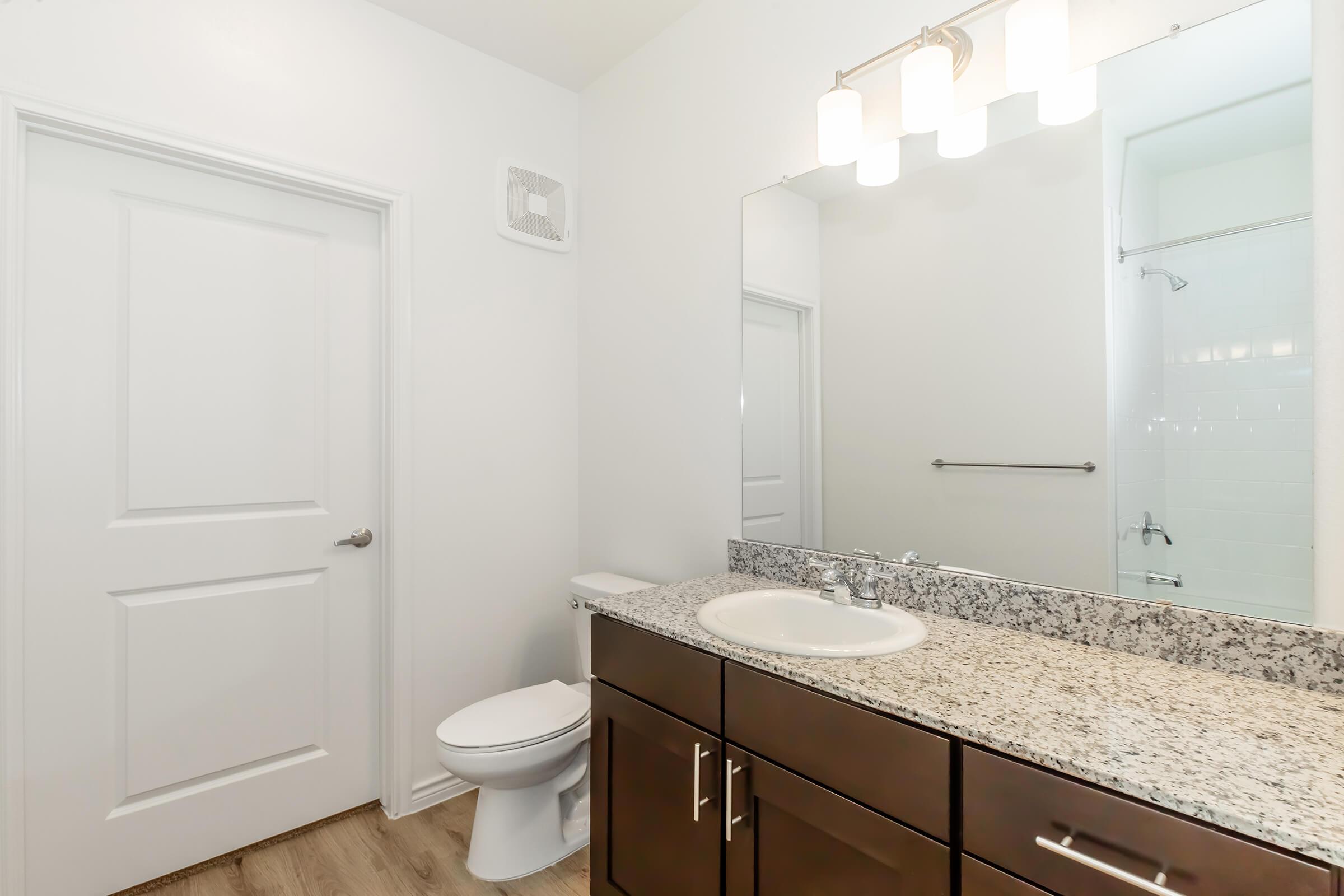 A clean and modern bathroom featuring a granite countertop with a sink, a wooden cabinet below, and a large mirror above. There is a white toilet next to the door, and a shower area is visible in the background. The walls are painted white, and the floor is made of light-colored wood.
