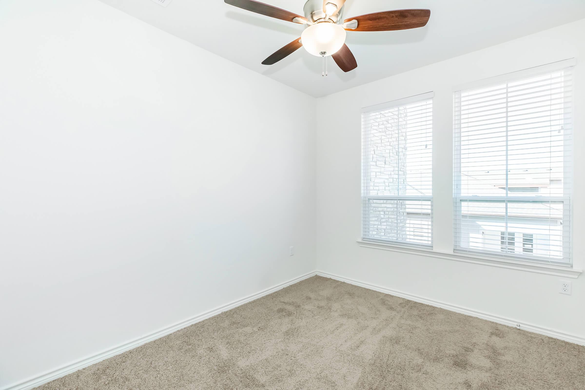 Empty room with light-colored walls, a ceiling fan with wooden blades, and two windows covered with blinds. The floor is covered with beige carpet, and the overall space is bright and minimalist.