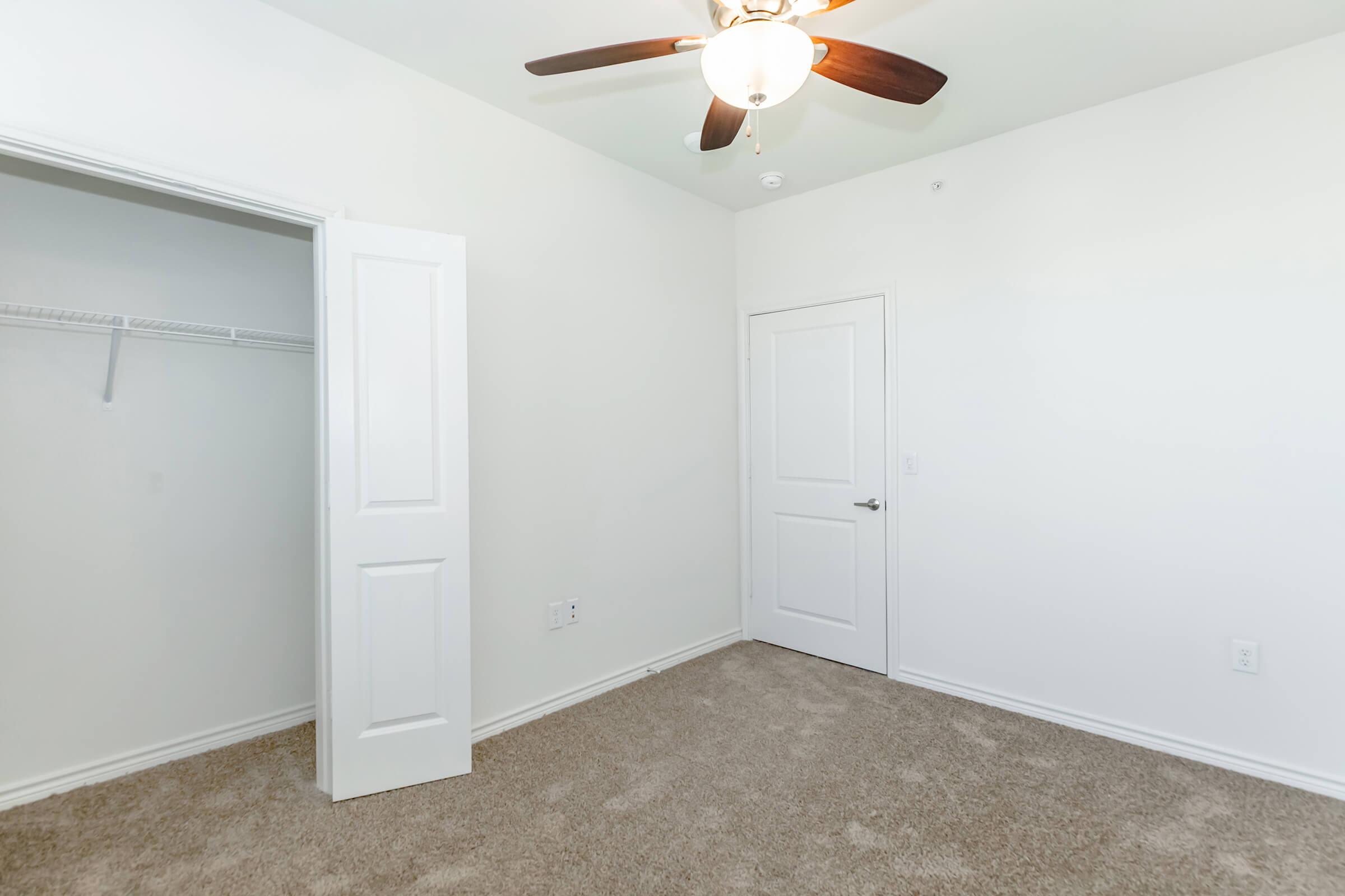 A bright, empty room featuring light-colored walls, a ceiling fan with wooden blades, and plush carpet. A closet with an open door is visible on the left side, while another door is on the right, leading to a different space. The room is well-lit and showcases a simple, modern design.