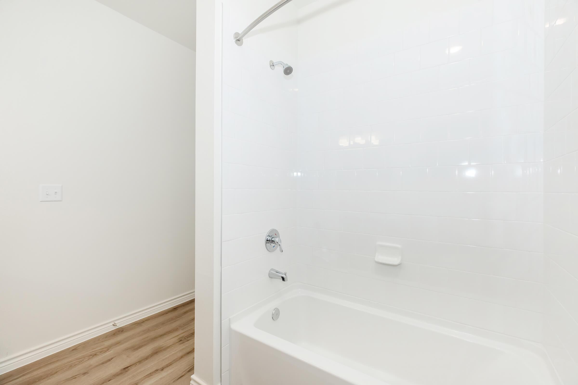 A clean, modern bathroom featuring a white tiled shower area with a silver showerhead and a bathtub. The wall is painted a light color, and the floor is covered with light wood-like flooring. Natural light illuminates the space, giving it a fresh and airy feel.