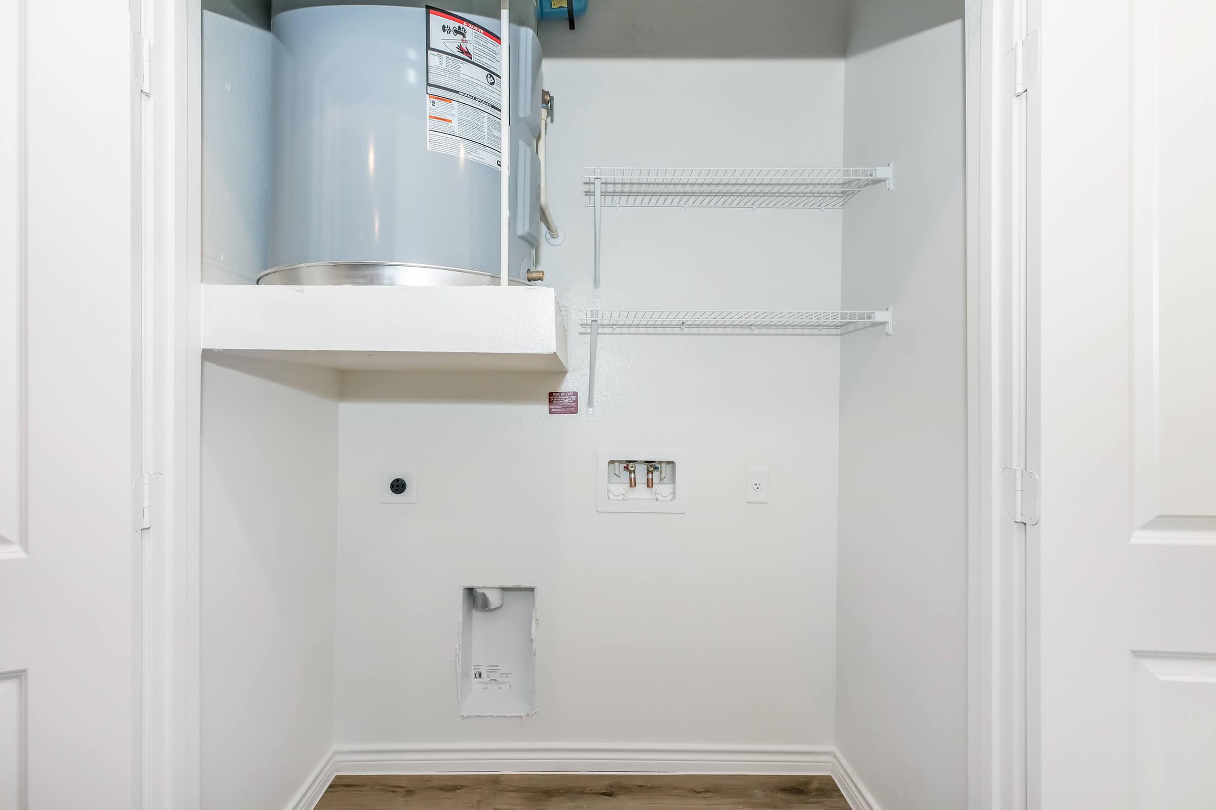 A small closet featuring a water heater mounted on the wall. There are two wire shelves for storage, and the wall beneath a utility panel shows a cutout. The floor is a light wooden laminate, and the walls are painted white, giving the space a clean and minimalistic appearance.