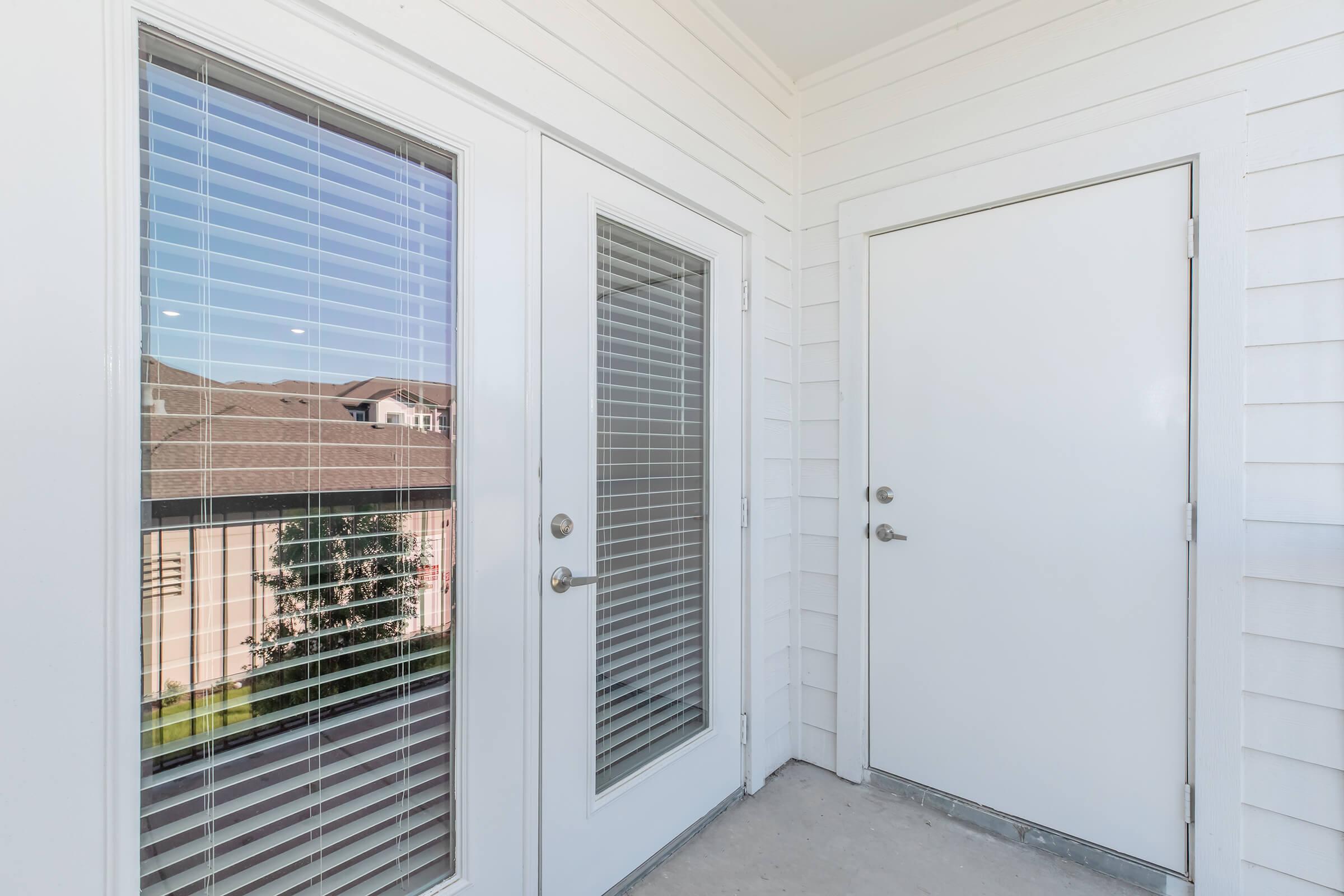 A corner view of a balcony featuring two glass doors with horizontal blinds on the left and a solid white door on the right. The balcony has a light-colored, textured wall, and the background shows rooftops and greenery outside.