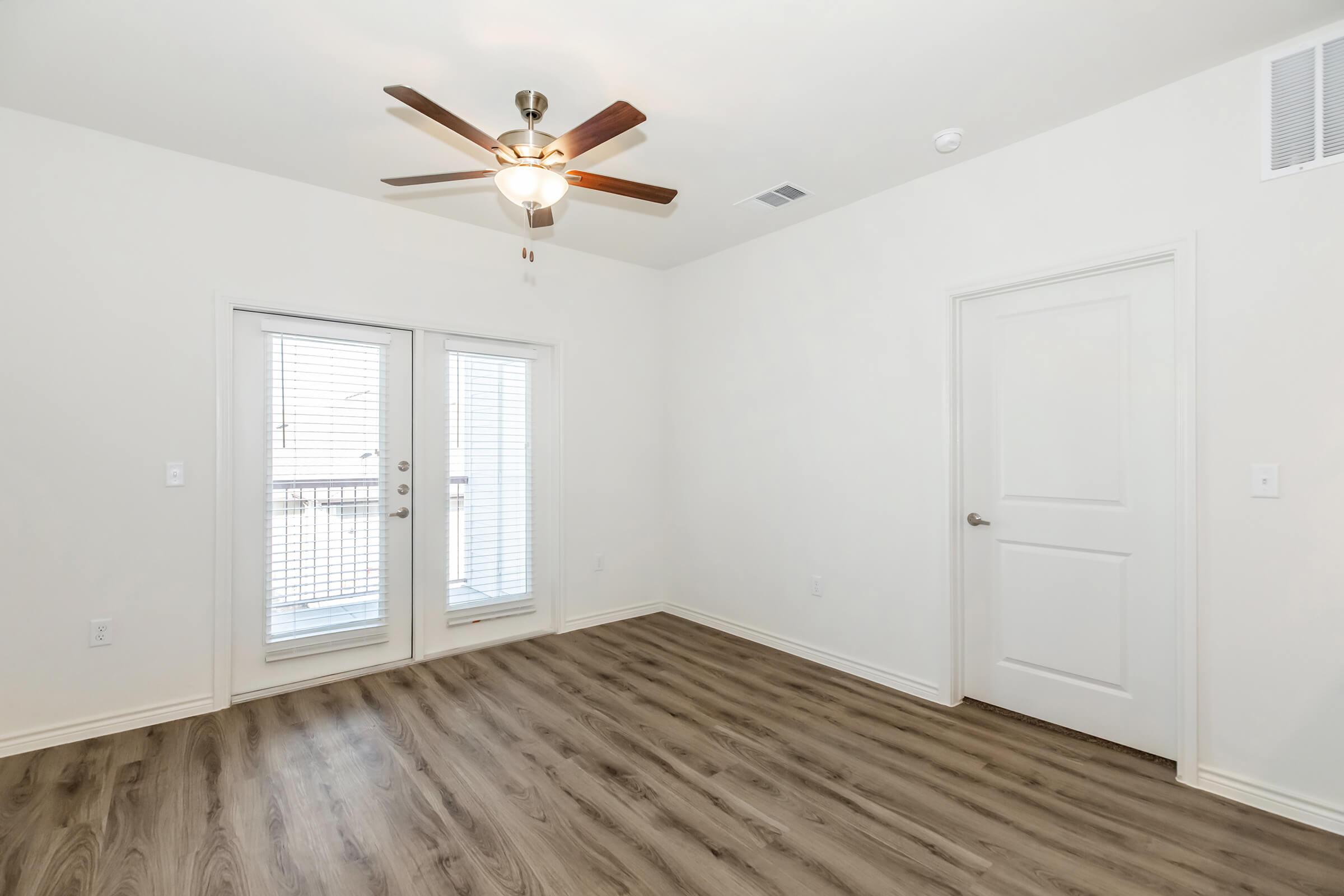 Empty room with light brown wooden flooring, a ceiling fan with light, and white walls. Large double doors lead to a balcony, and a single door on the right likely leads to another room. Natural light enters through the windowed doors, creating a bright, airy atmosphere.
