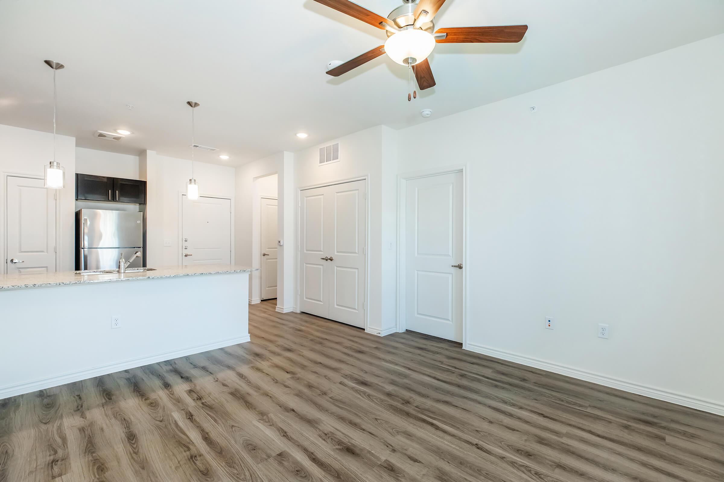 Interior view of a modern apartment featuring a spacious living area with laminate flooring, a ceiling fan, and an open kitchen with a granite countertop. The kitchen includes pendant lighting and stainless steel appliances. There are white walls and doors, enhancing the bright and airy atmosphere.