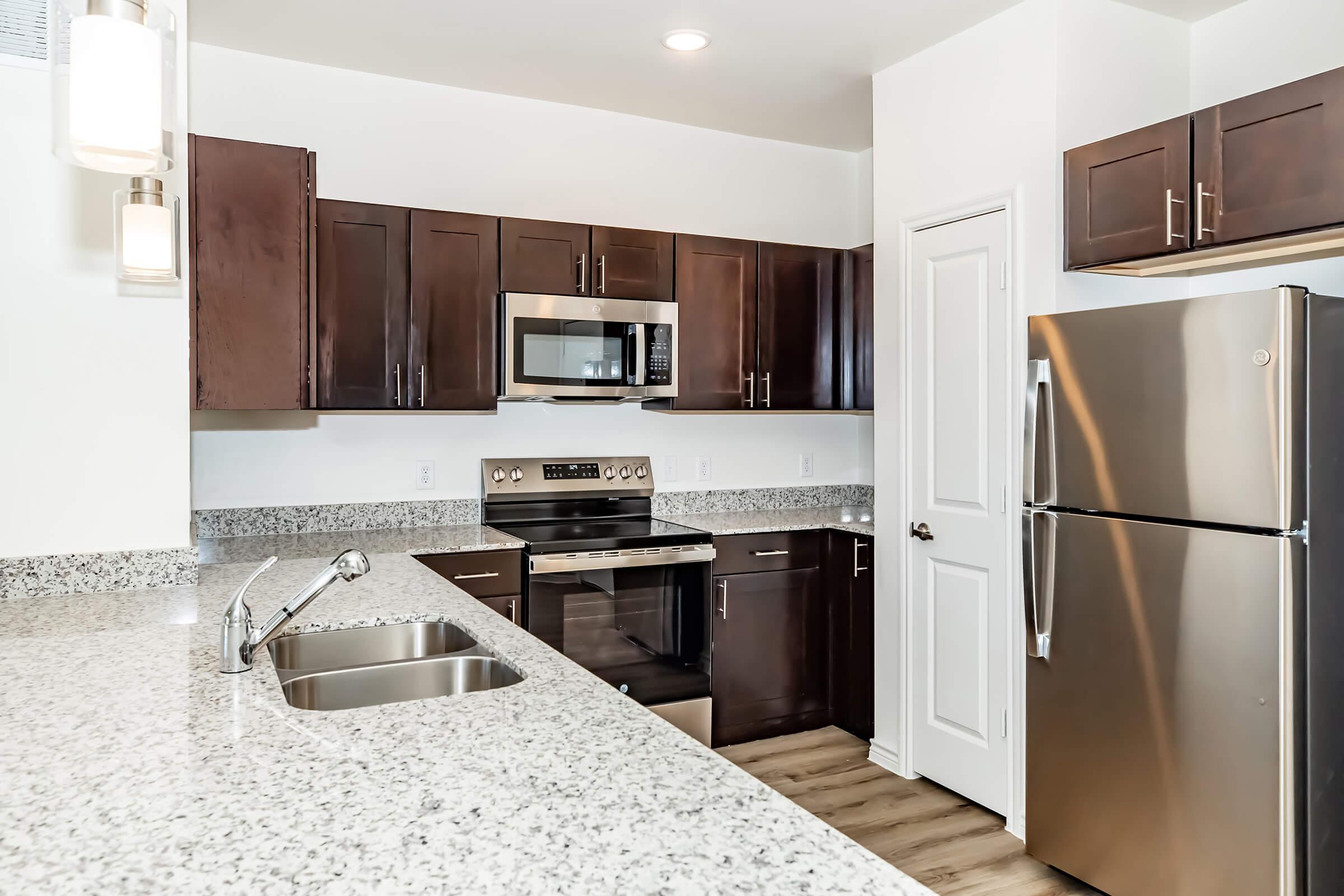 Modern kitchen featuring dark wood cabinets, stainless steel appliances including a microwave and refrigerator, and a granite countertop. The space is well-lit with pendant lighting and has a sleek, contemporary design.