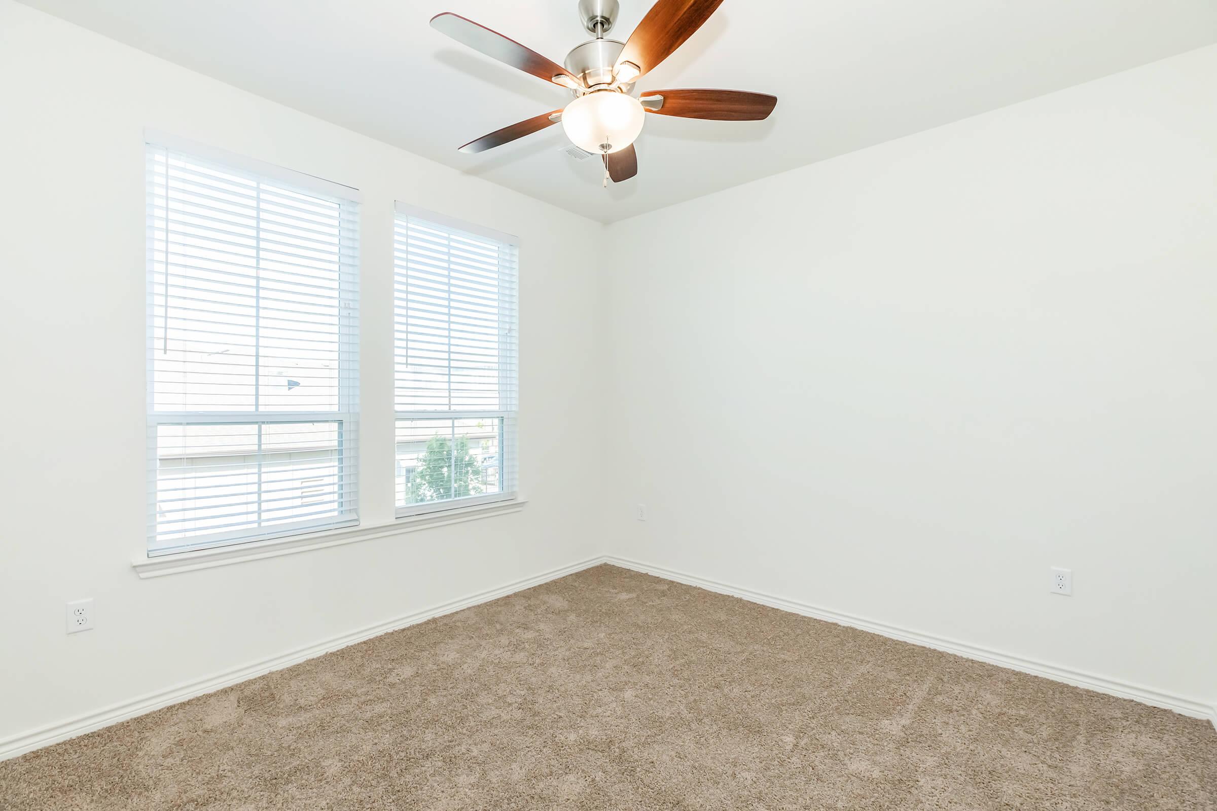 Empty room with light brown carpet, white walls, and ceiling fan. Two windows with blinds allow natural light to enter, creating a bright and airy atmosphere. The space is uncluttered and ready for furnishing.