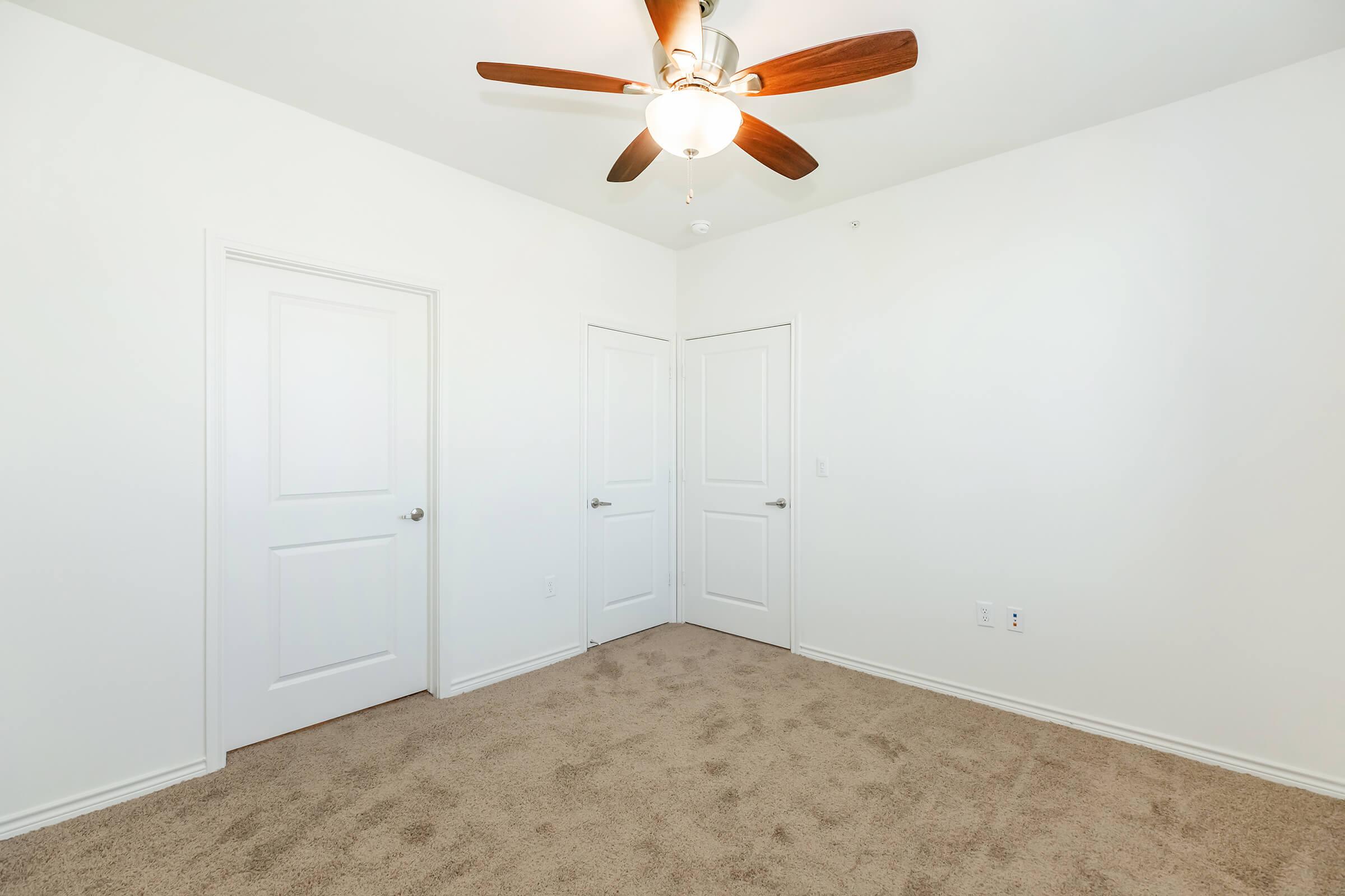 A spacious, empty room featuring light beige carpet and white walls. There are two closed doors on the right side of the image and a ceiling fan with wooden blades in the center. The room appears bright and well-lit, with a simple and clean design.