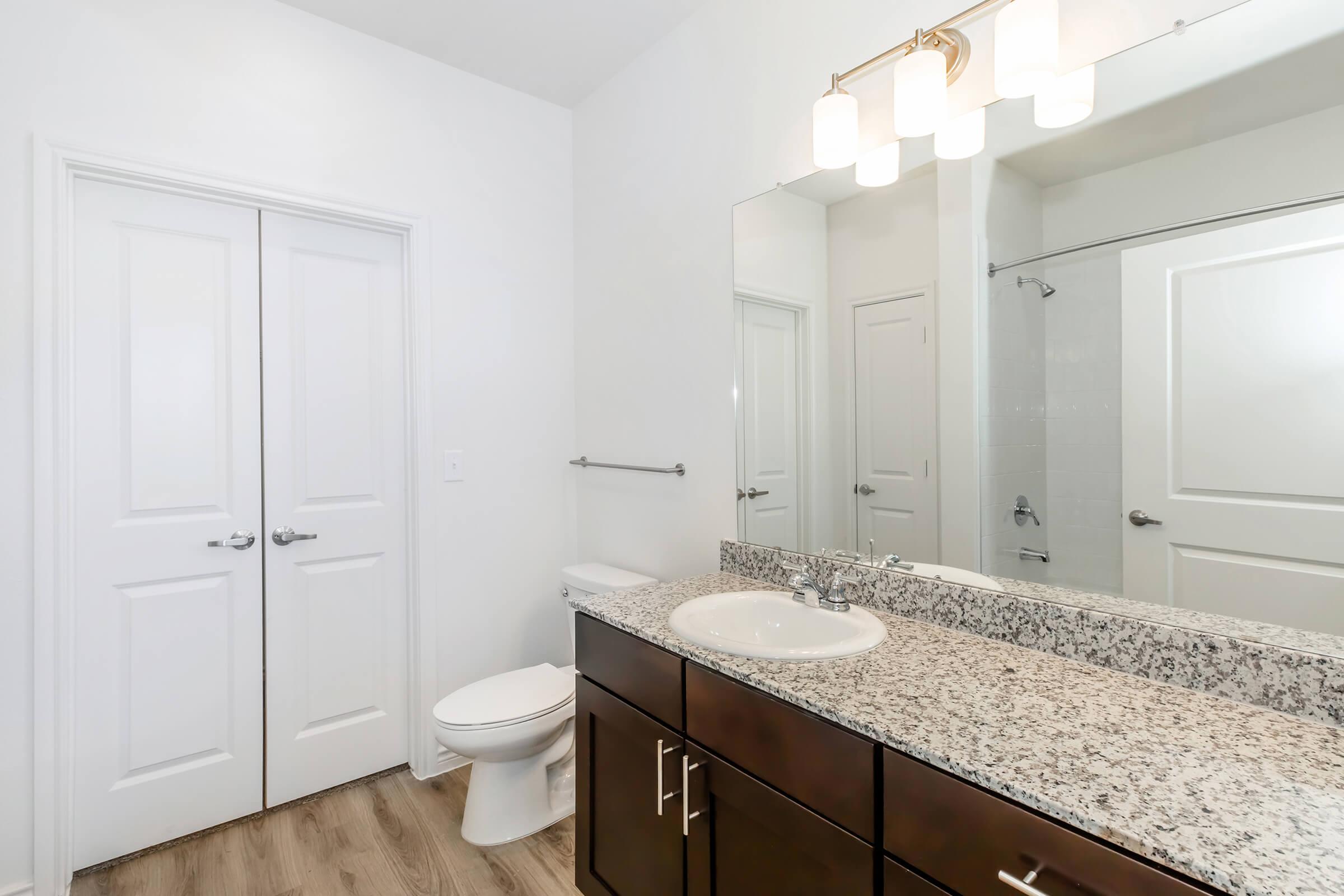 Modern bathroom featuring a double-door closet, a white toilet, and a granite countertop with a sink. Above the sink, there is a stylish light fixture. The walls are painted white, and a glass shower enclosure is visible in the background, creating a clean and contemporary look.