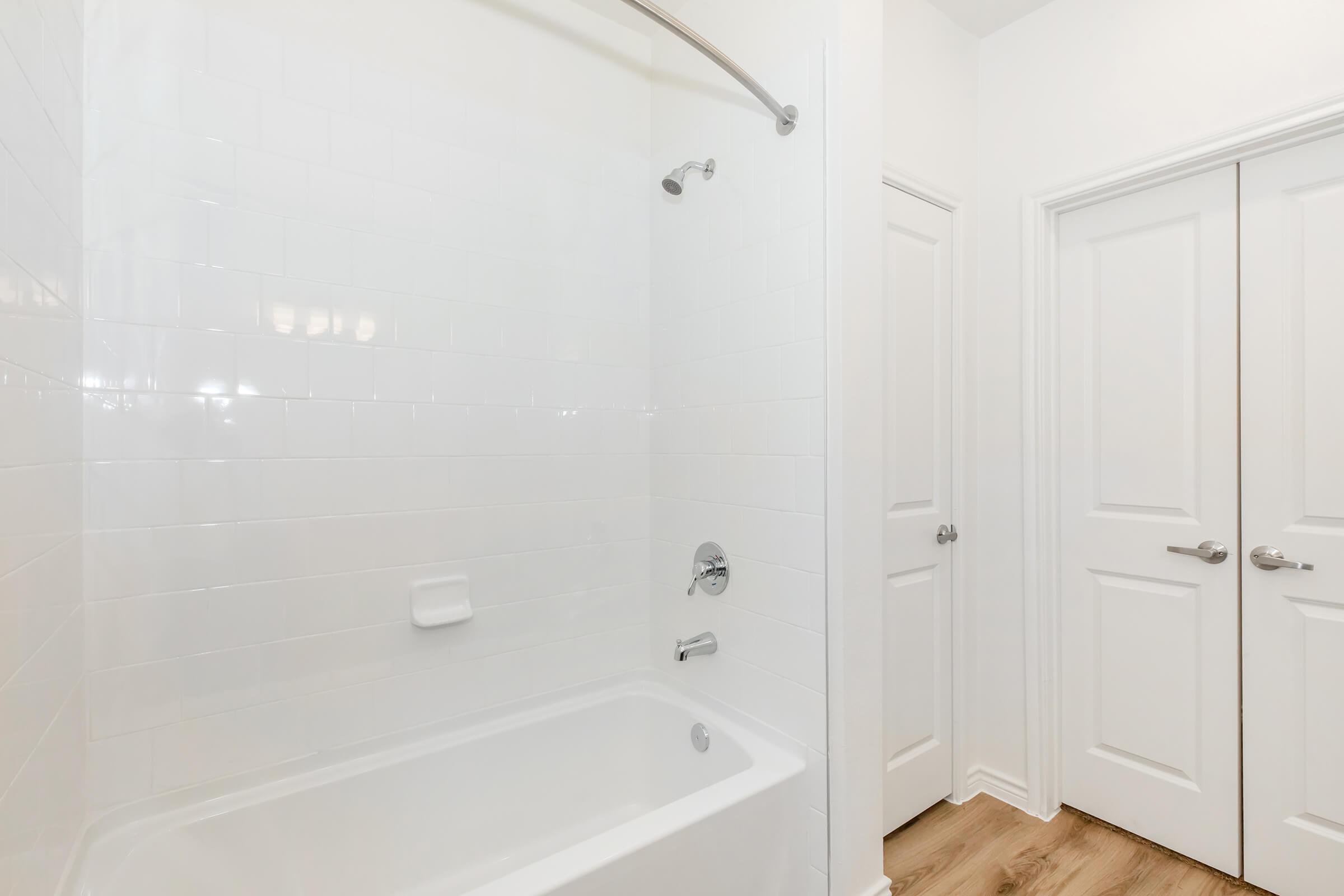 Clean bathroom with a white tiled shower and bathtub. The space features a curved shower rod, a single faucet, and a small shelf. Light hardwood flooring complements the bright, minimalist design, with two closed doors visible in the background.