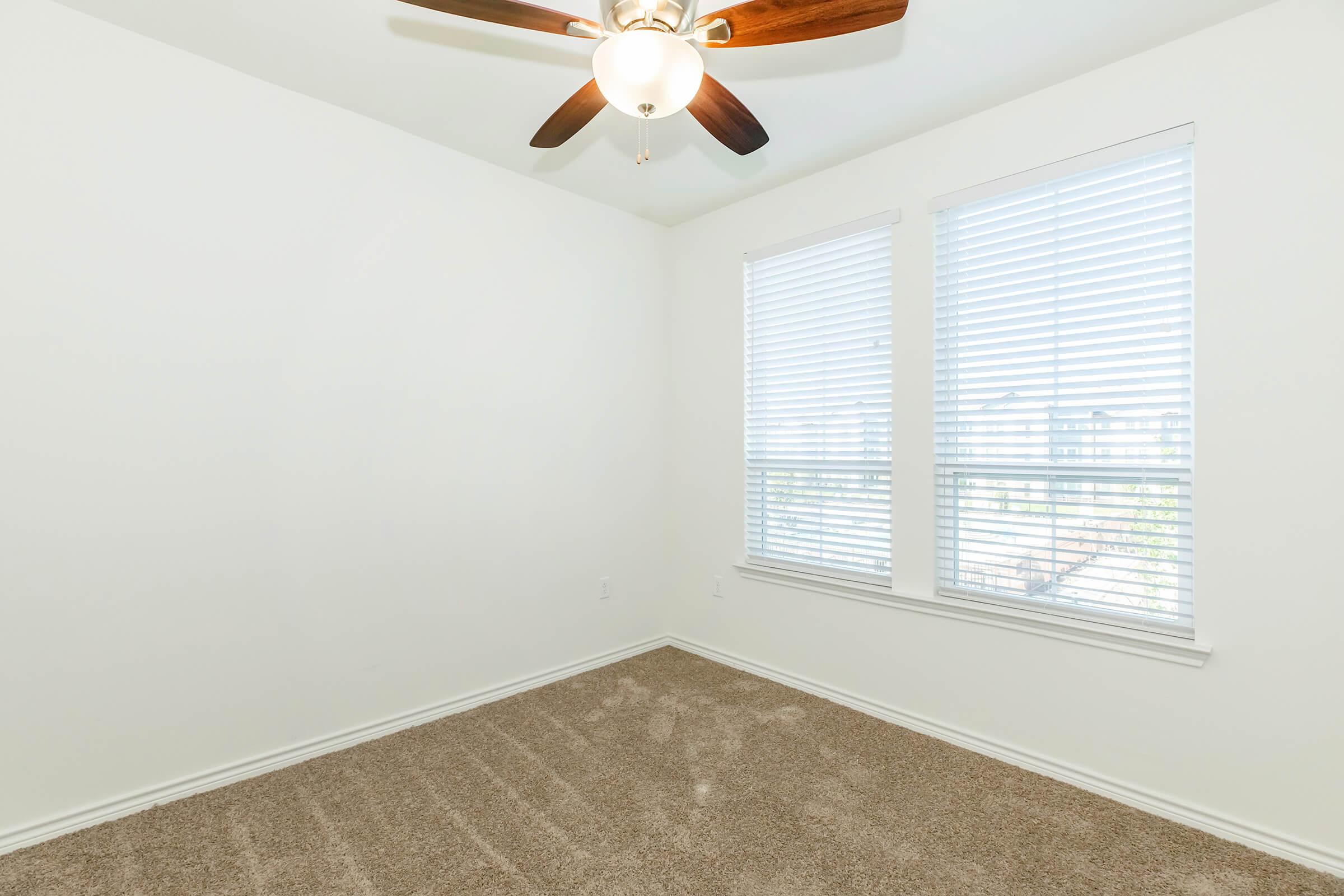Empty room with light-colored walls and a ceiling fan. Two windows with white blinds allow natural light in. The floor is covered with plush, beige carpeting. The space appears clean and ready for furnishing.