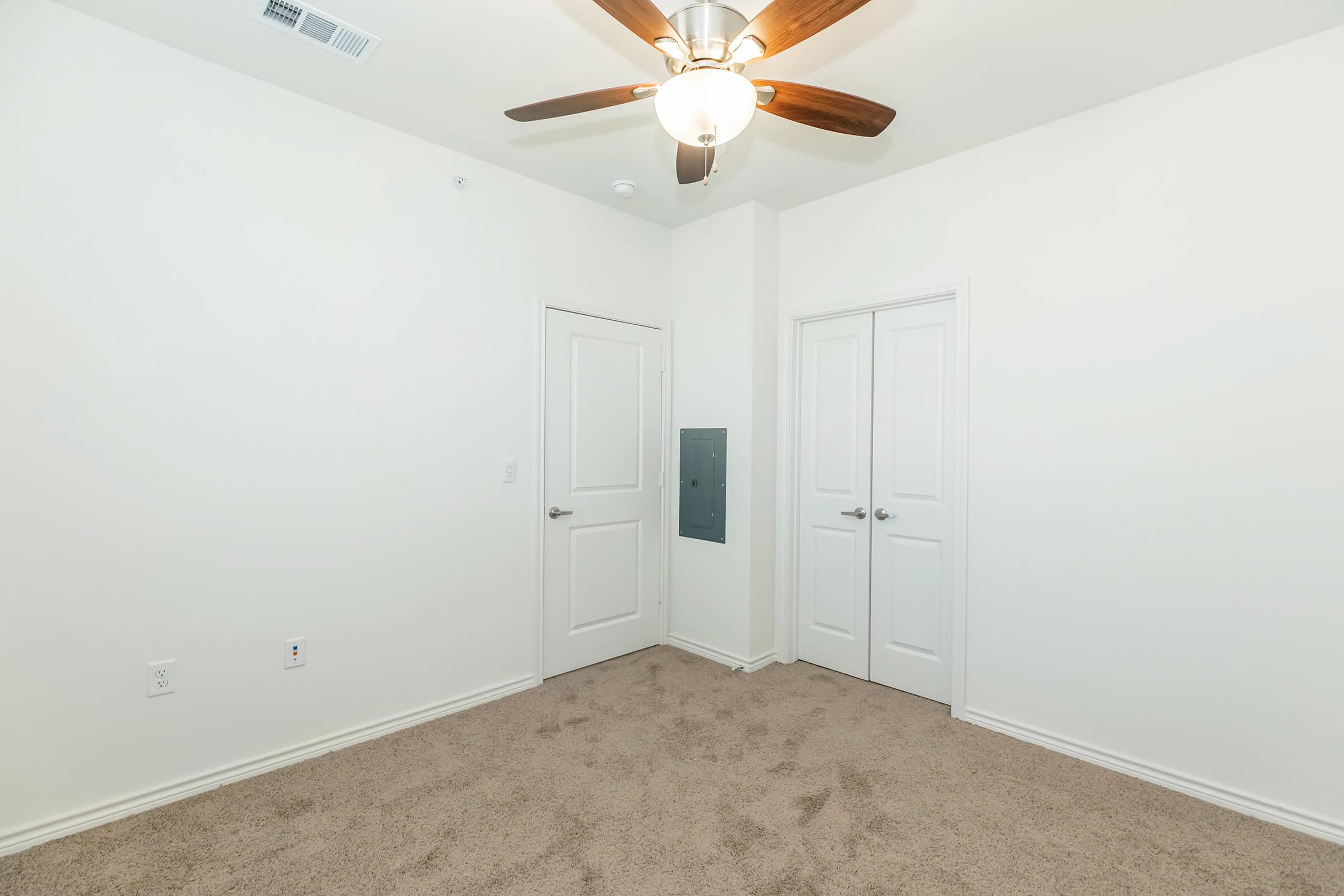 A bare room with light beige carpet and white walls. There is a ceiling fan with a light fixture, and two closed doors on the right side. The left side features a wall-mounted electrical panel. The room appears empty and well-lit, conveying a minimalistic aesthetic.