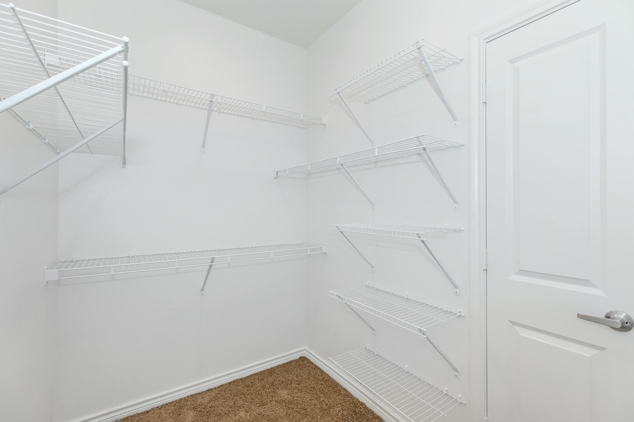 Empty closet with white wire shelving mounted on the walls, featuring several shelves arranged at varying heights. The walls are painted white, and the floor is covered with beige carpet. A door is visible on the right side of the image.