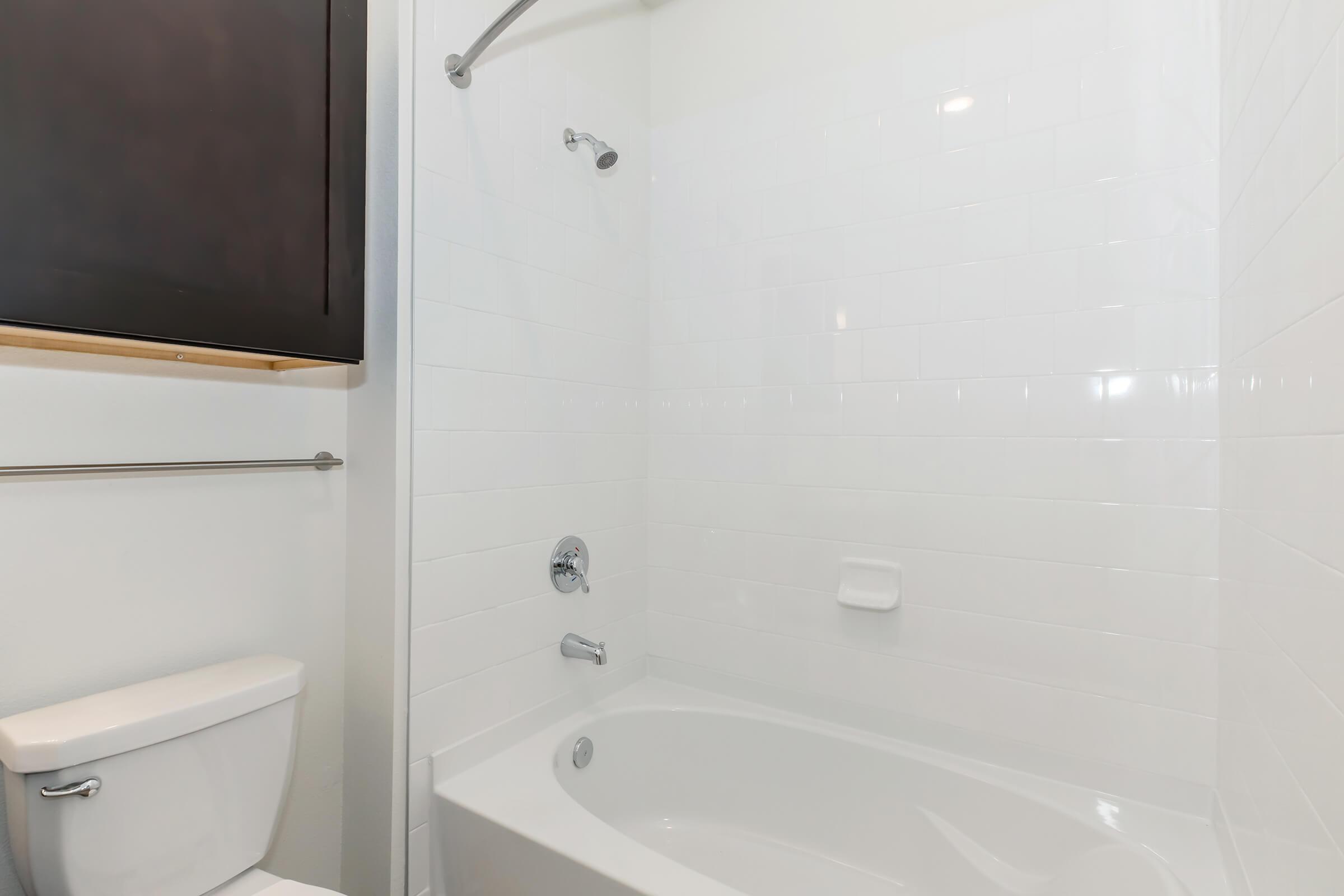 A clean, modern bathroom featuring a white bathtub and shower combination, a wall-mounted faucet, and a toilet. The walls are tiled in glossy white, with a sleek dark cabinet above the tub area. The overall design is minimalist and bright, creating an inviting space.