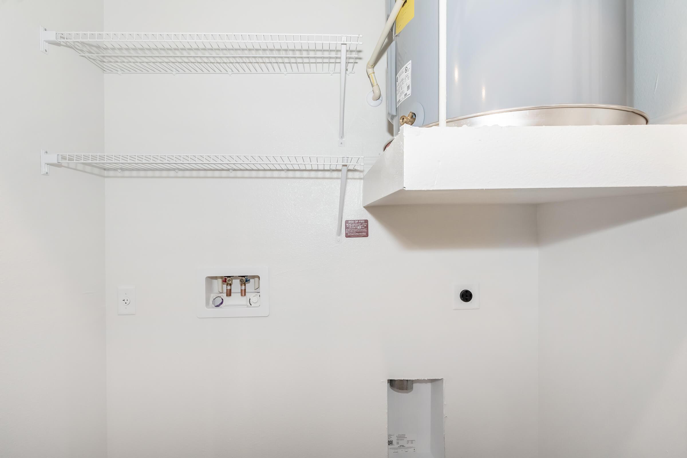 A clean, white utility room featuring a mounted water heater, two metal wire shelves on the left wall, and a small panel with plumbing connections. There is a hole in the bottom right wall, and power outlets are visible, providing a functional and organized space for maintenance access.