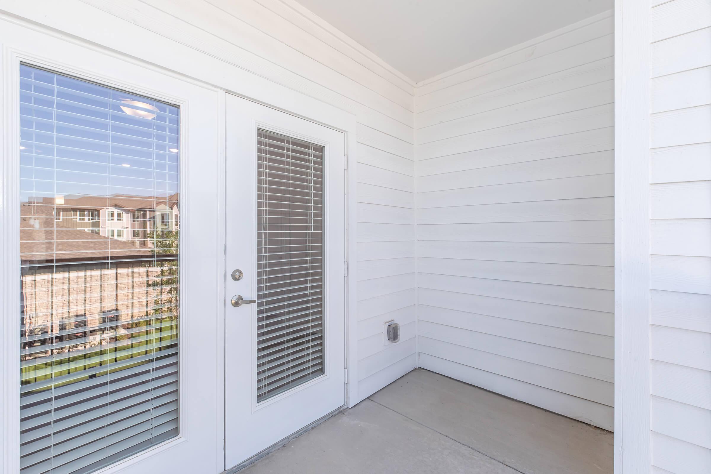 An empty balcony with white walls and large sliding glass doors featuring blinds. Natural light streams in, highlighting the clean, modern design. The view outside includes residential buildings, creating a serene outdoor space. The floor is concrete, providing a minimalist aesthetic.